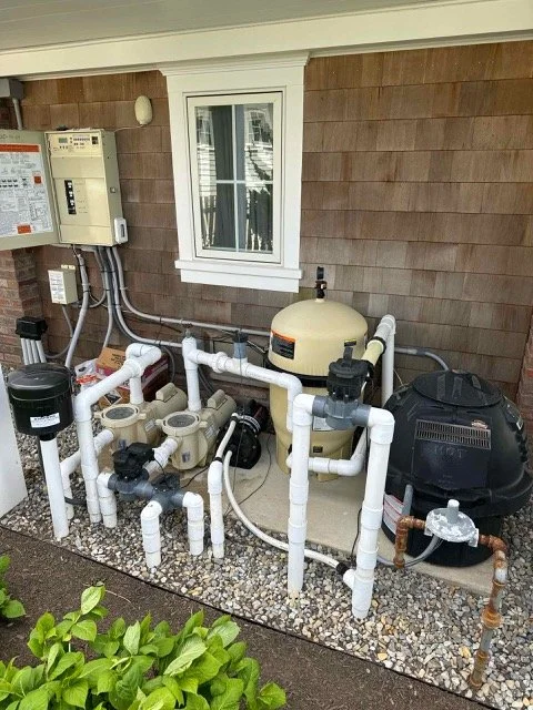 Outdoor pool equipment setup with white PVC pipes, a pool filter, and a control panel on the house wall.