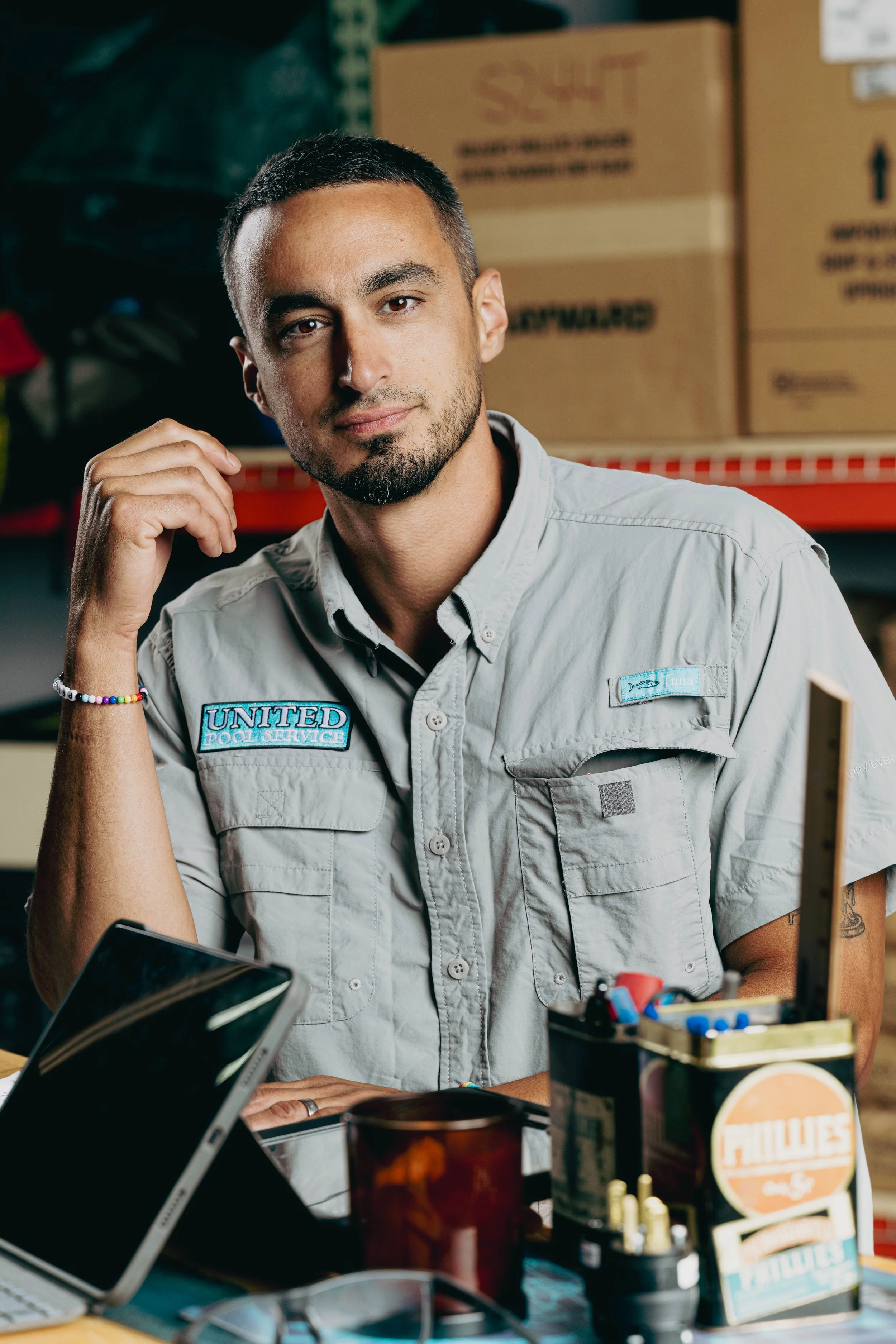 A man with short dark hair and a beard, wearing a light gray uniform with 'United Pool Service' patches, sitting at a cluttered desk in a warehouse with boxes in the background.