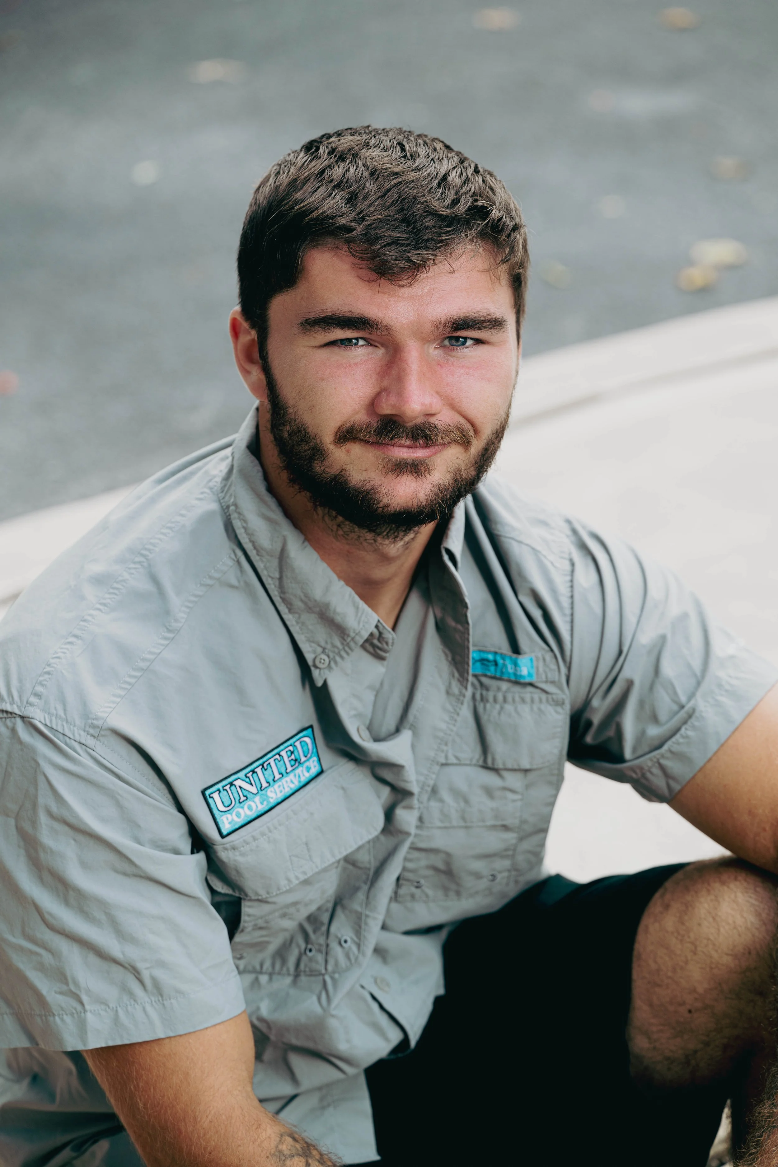A man with a beard and short brown hair, wearing a beige shirt with 'United Pool Service' patches, sitting outdoors on a concrete surface.