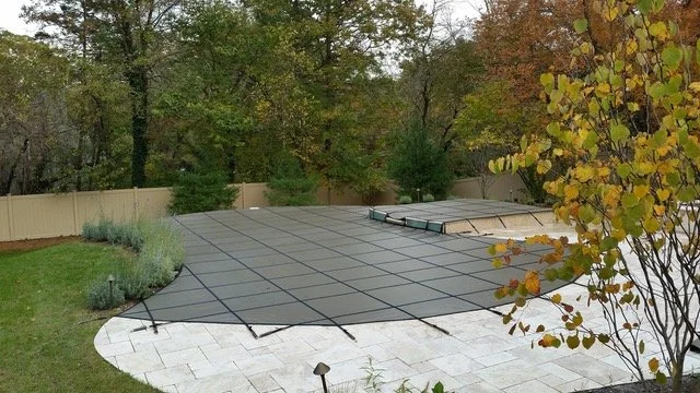 Swimming pool with cover on a patio, surrounded by a garden with trees and a beige fence.