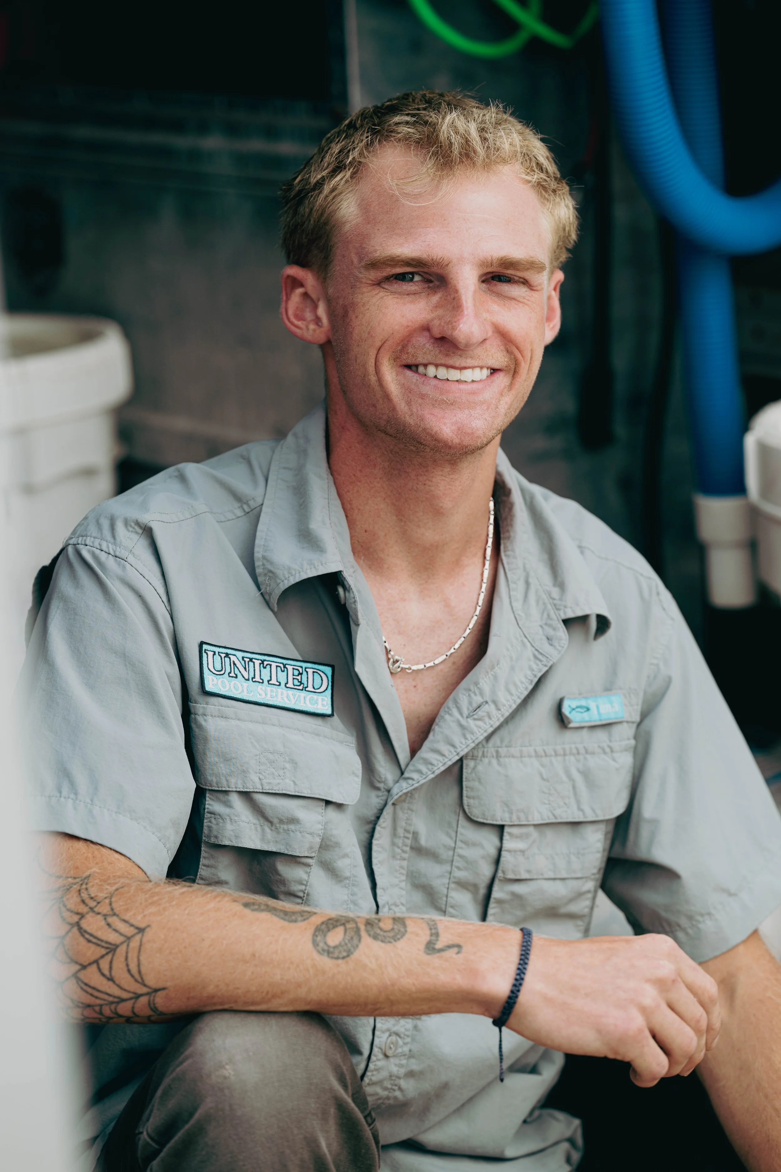 A smiling man in a gray uniform with a 'United Pool Service' patch, sitting in what appears to be a utility room with blue and green hoses in the background.