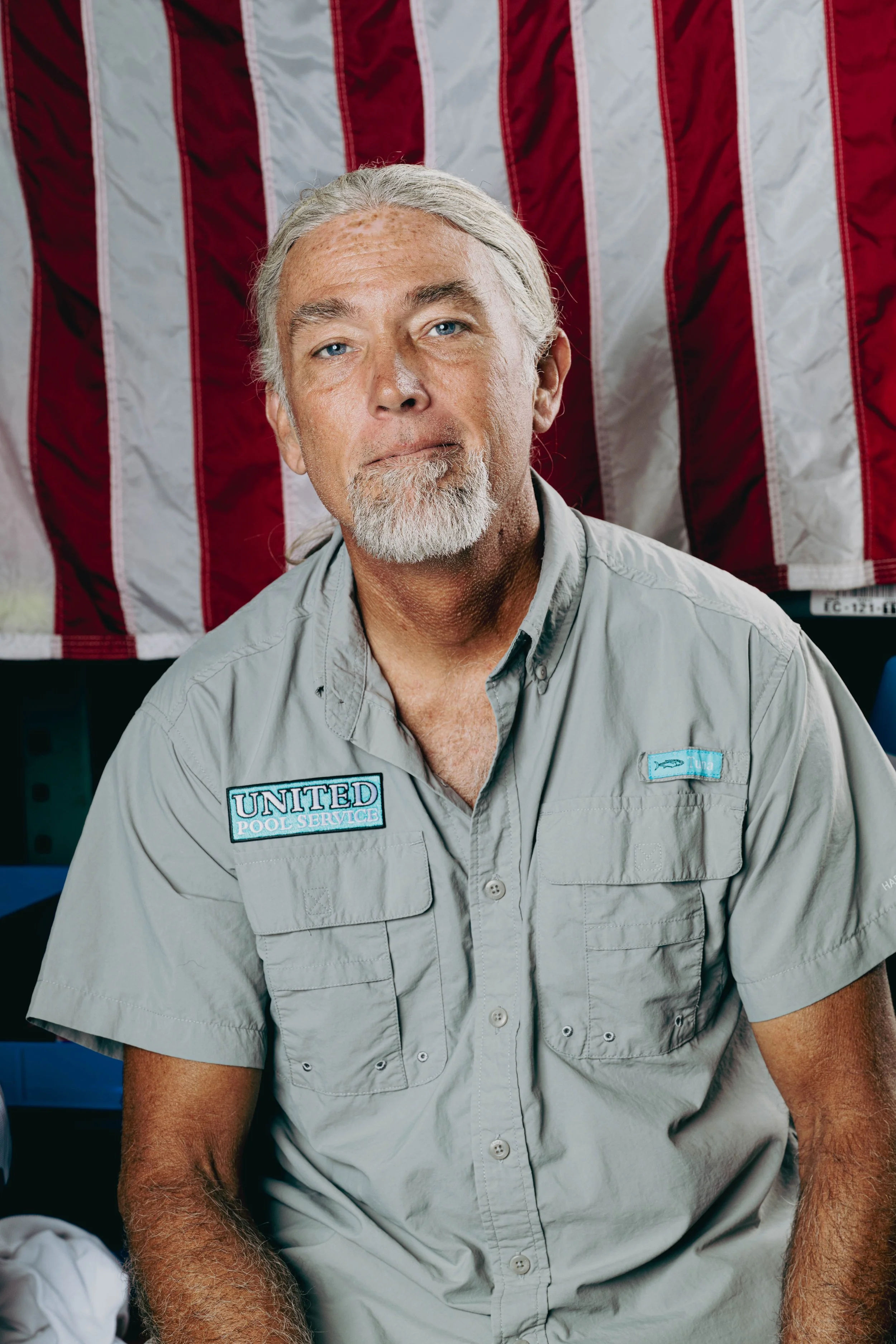 A man with gray hair and a beard wearing a uniform with patches for United Pool Service, sitting in front of an American flag.