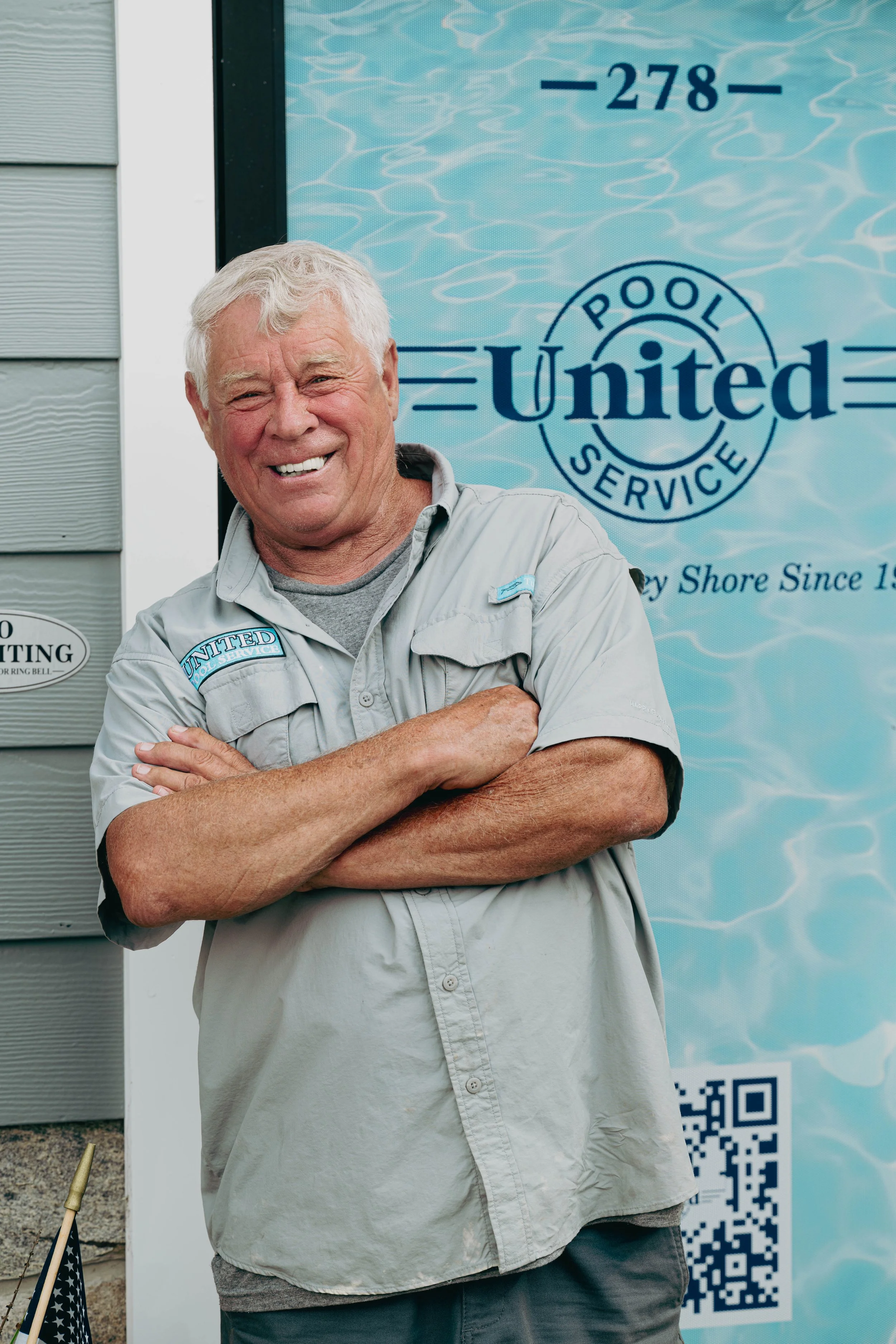 A smiling older man with gray hair and a crew cut, wearing a light gray work shirt with "United" patches, standing with arms crossed in front of a blue sign that reads 'Pool Service United' and displays a QR code.