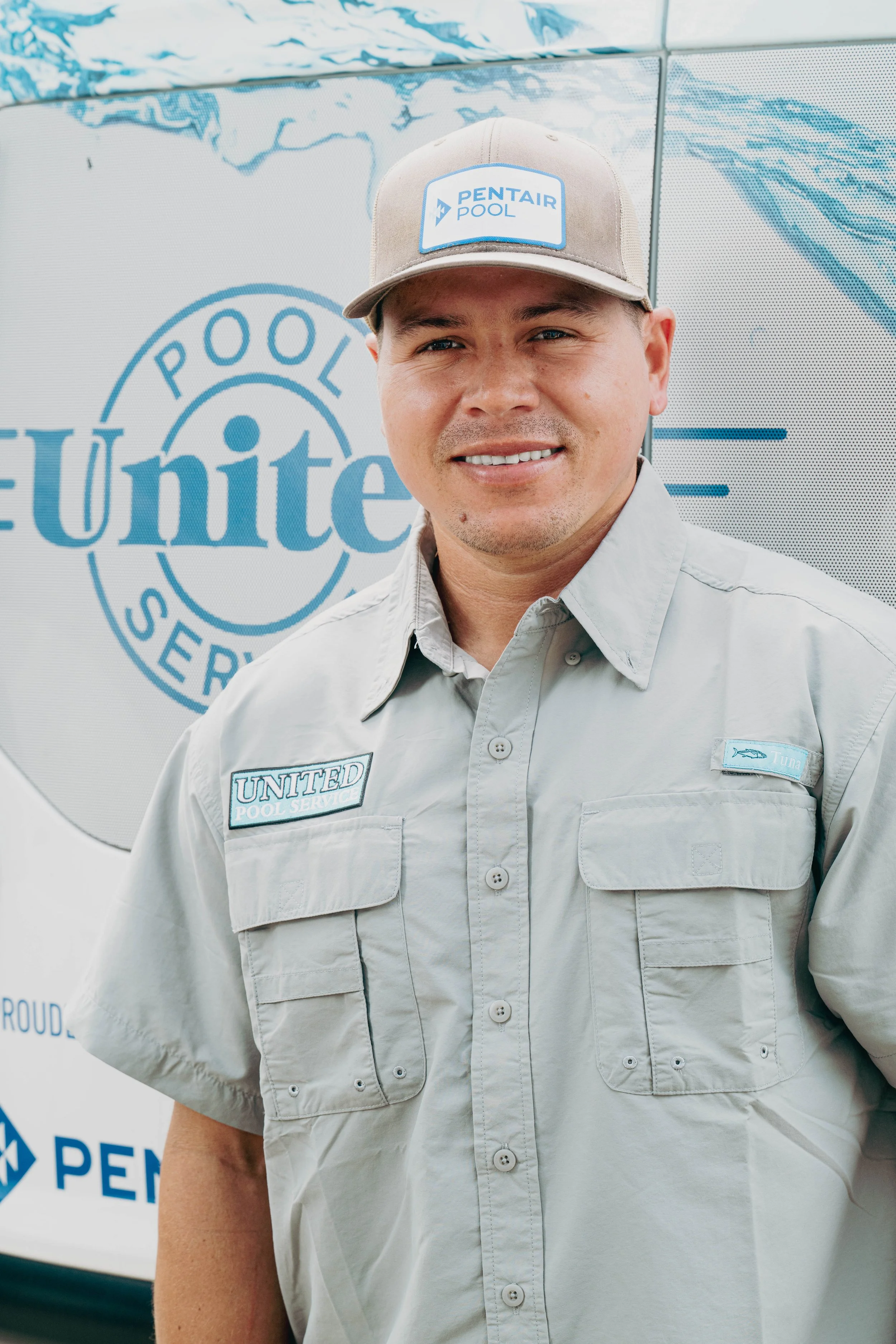A man wearing a beige uniform with logos and patches, standing in front of a large sign that reads 'Pool Service United'. He is also wearing a beige cap with a logo that reads 'Pentair Pool'.