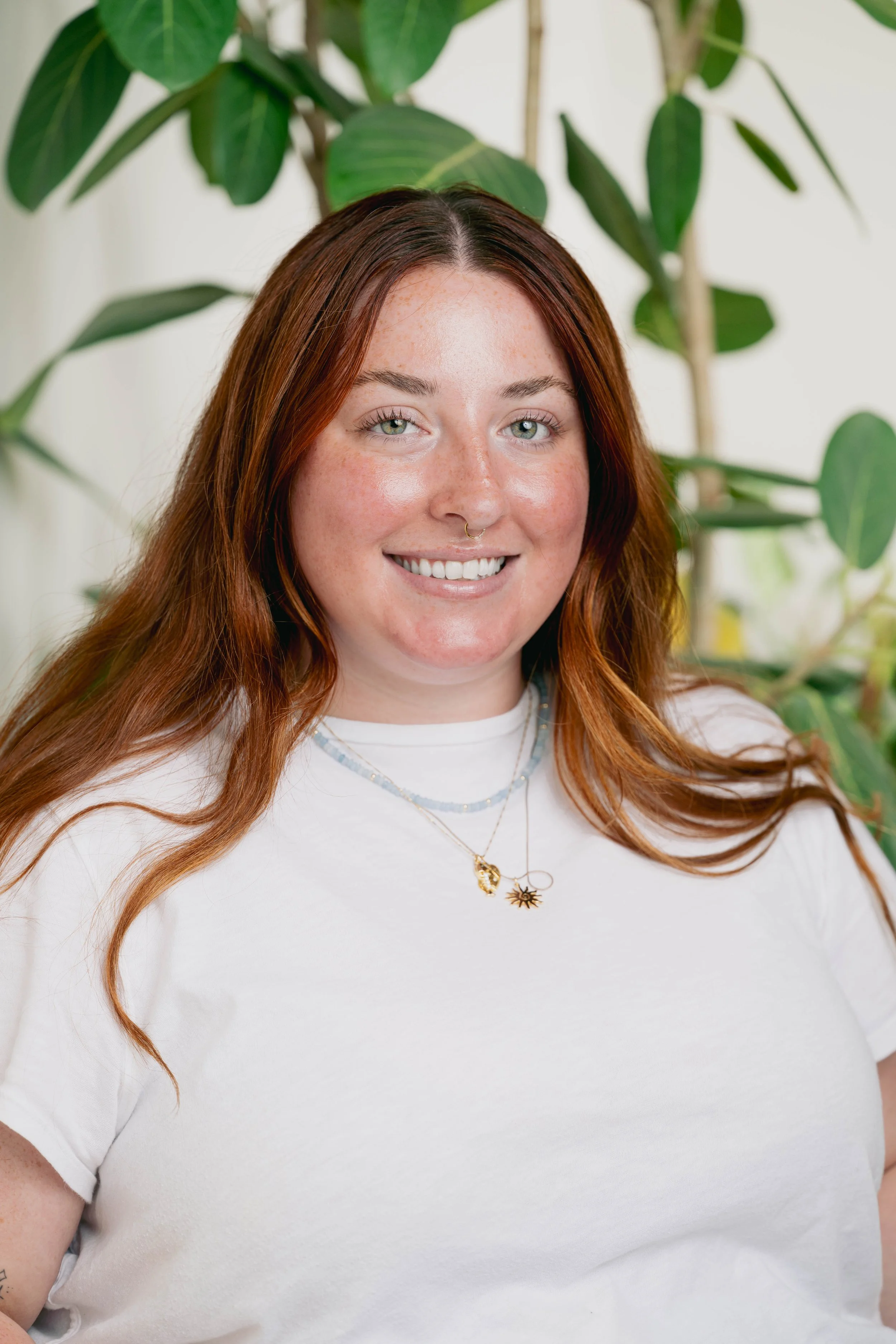 A woman with long, wavy red hair and light green eyes, smiling and wearing a white t-shirt with layered necklaces, standing in front of green leafy plants.