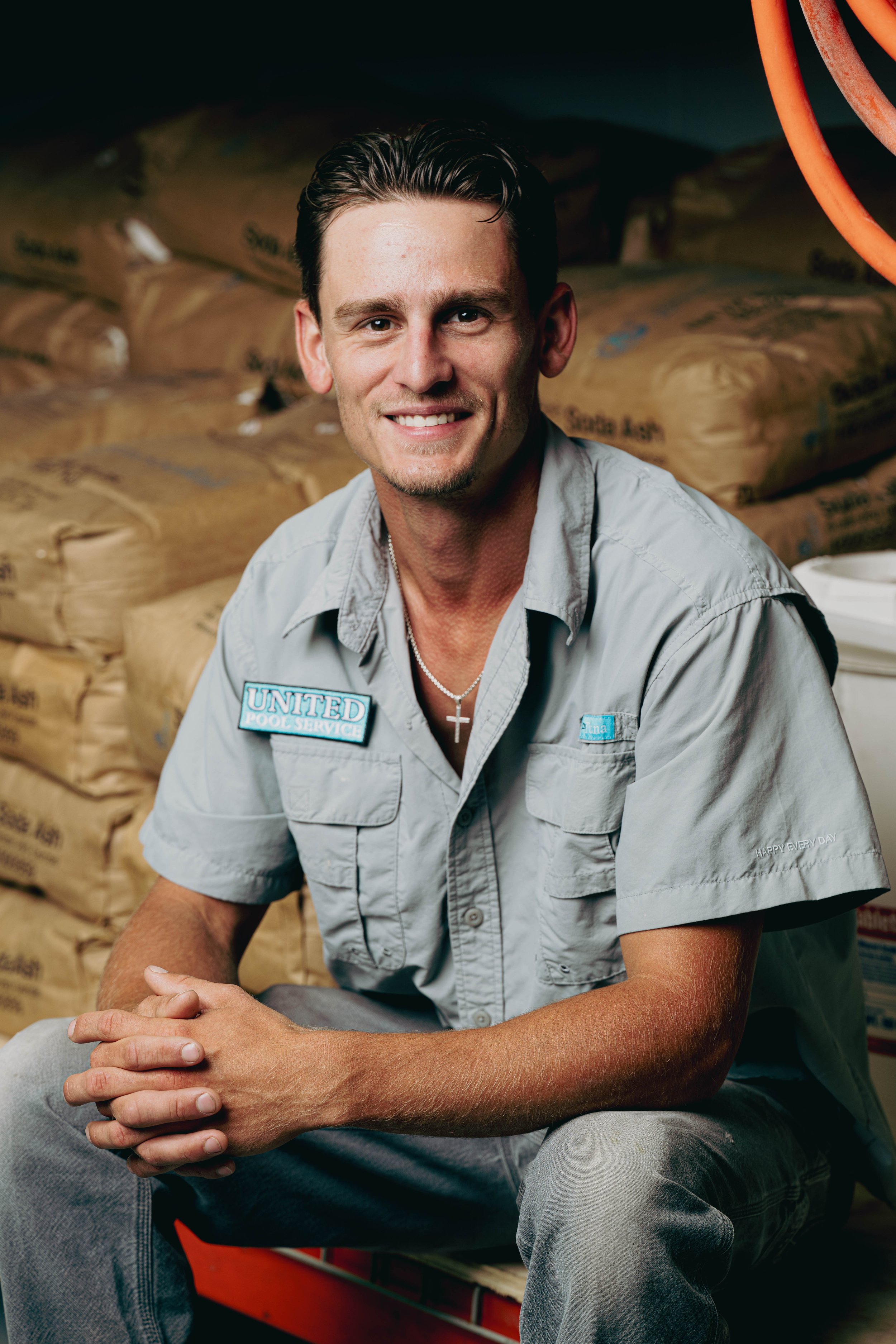 A smiling man in a gray uniform with a 'United Pool Service' patch on the shirt, sitting in front of stacked bags of pool salt.