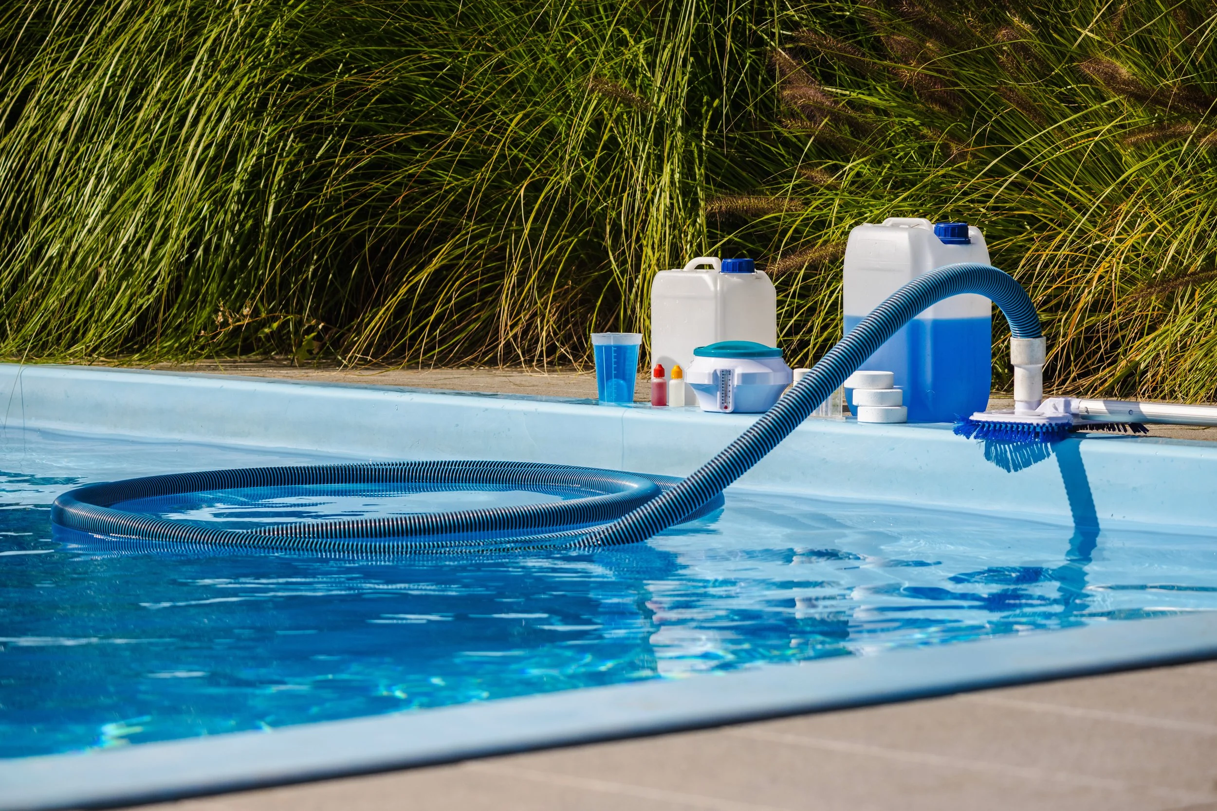 Poolside scene with cleaning supplies, including a pool vacuum hose, detergent bottles, a bucket, and a cup, next to a swimming pool with blue water and surrounded by green plants.