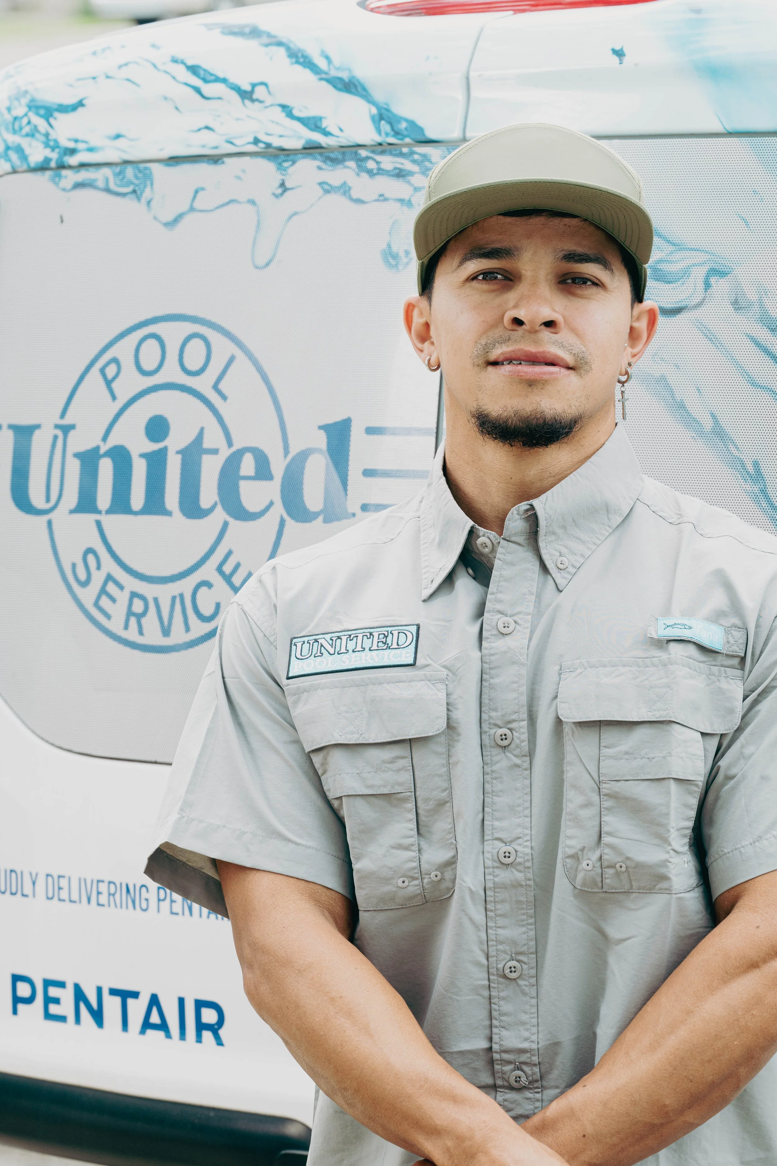 A pool service technician standing in front of a branded vehicle with the logo 'Pool United Service'. The technician is wearing a light gray uniform with patches, a beige cap, earrings, and has a goatee.