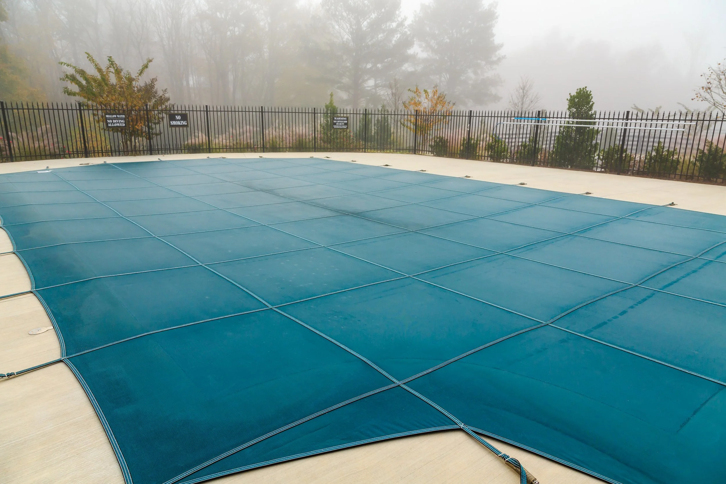 Empty outdoor swimming pool covered with a blue pool cover, surrounded by a black metal fence, with foggy trees in the background.