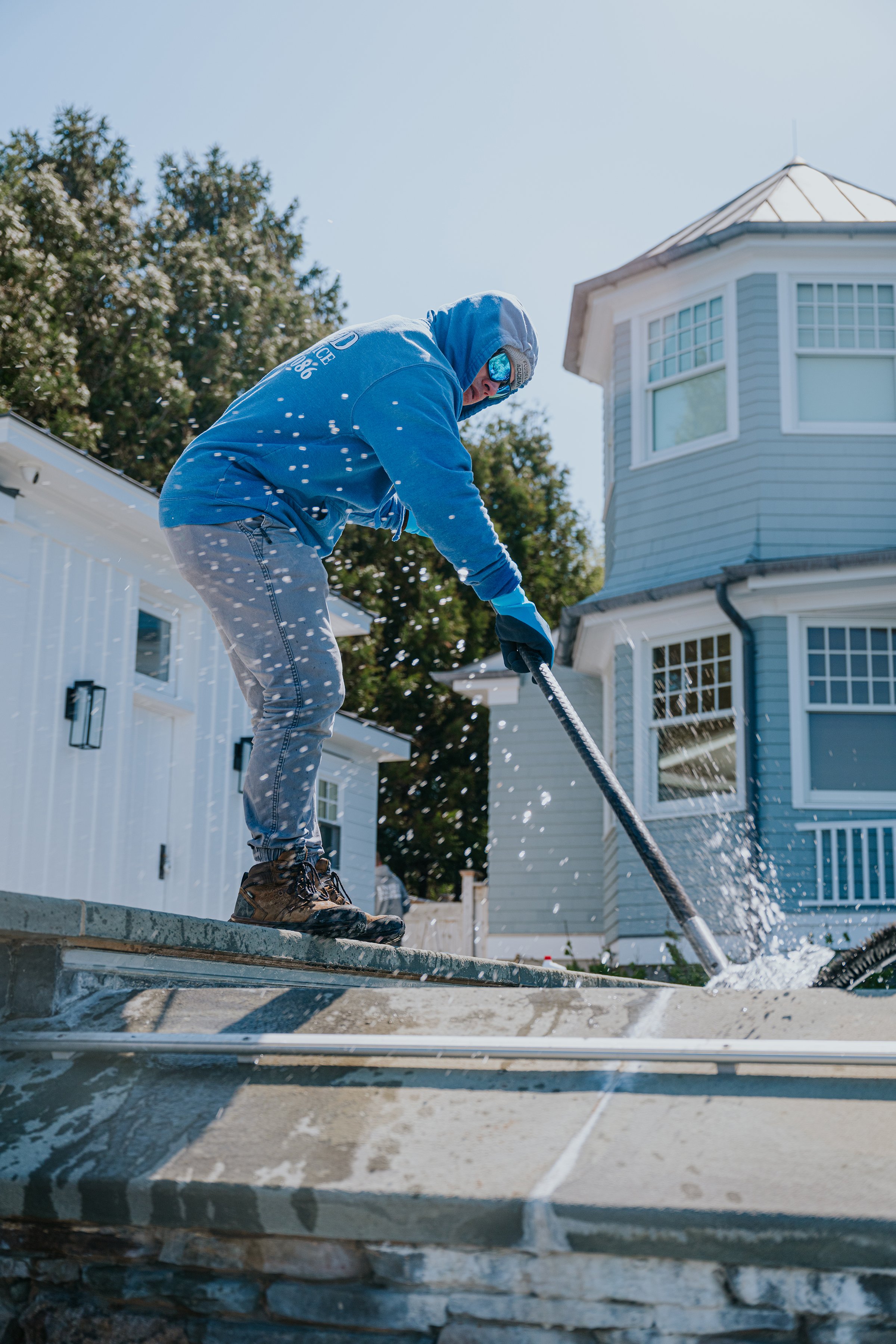 A worker in a blue hoodie, jeans, gloves, and sunglasses cleaning a stone pathway outside a house with a pressure washer on a sunny day.