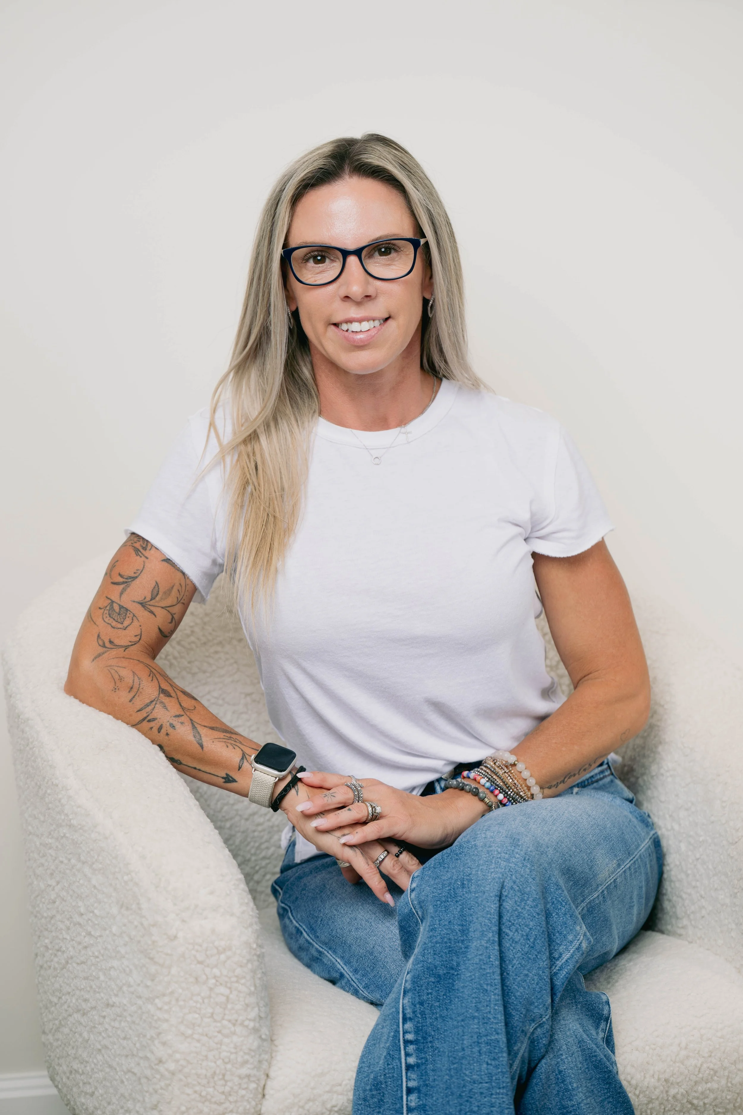 A woman with long blonde hair, wearing glasses, a white t-shirt, and jeans, sitting on a beige textured armchair, smiling at the camera.