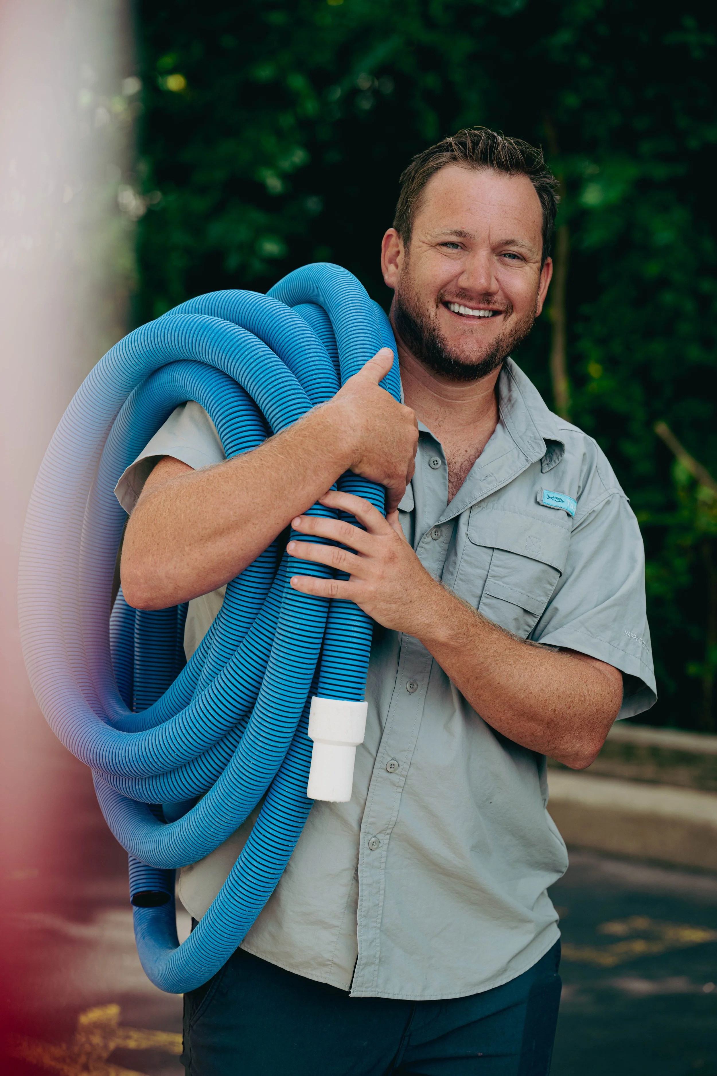 A smiling man in a light gray short-sleeve shirt is holding a coil of blue flexible tubing over his shoulder, standing outdoors with trees in the background.