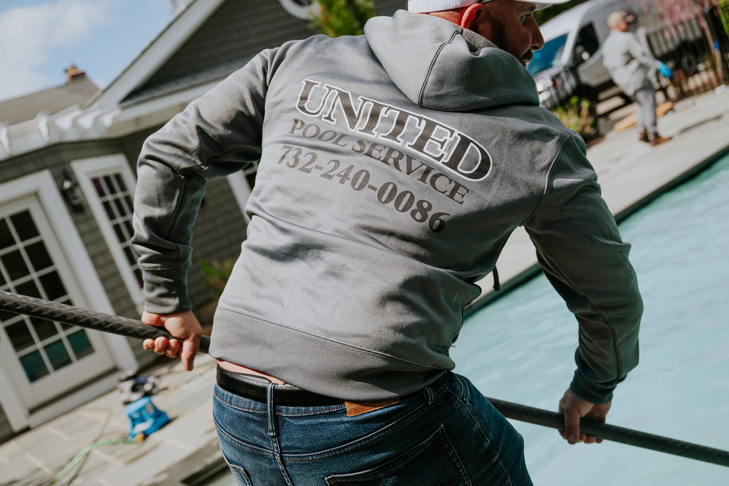 A man in a gray hoodie with 'United Pool Service' and a phone number on the back, working near a swimming pool with a hose.