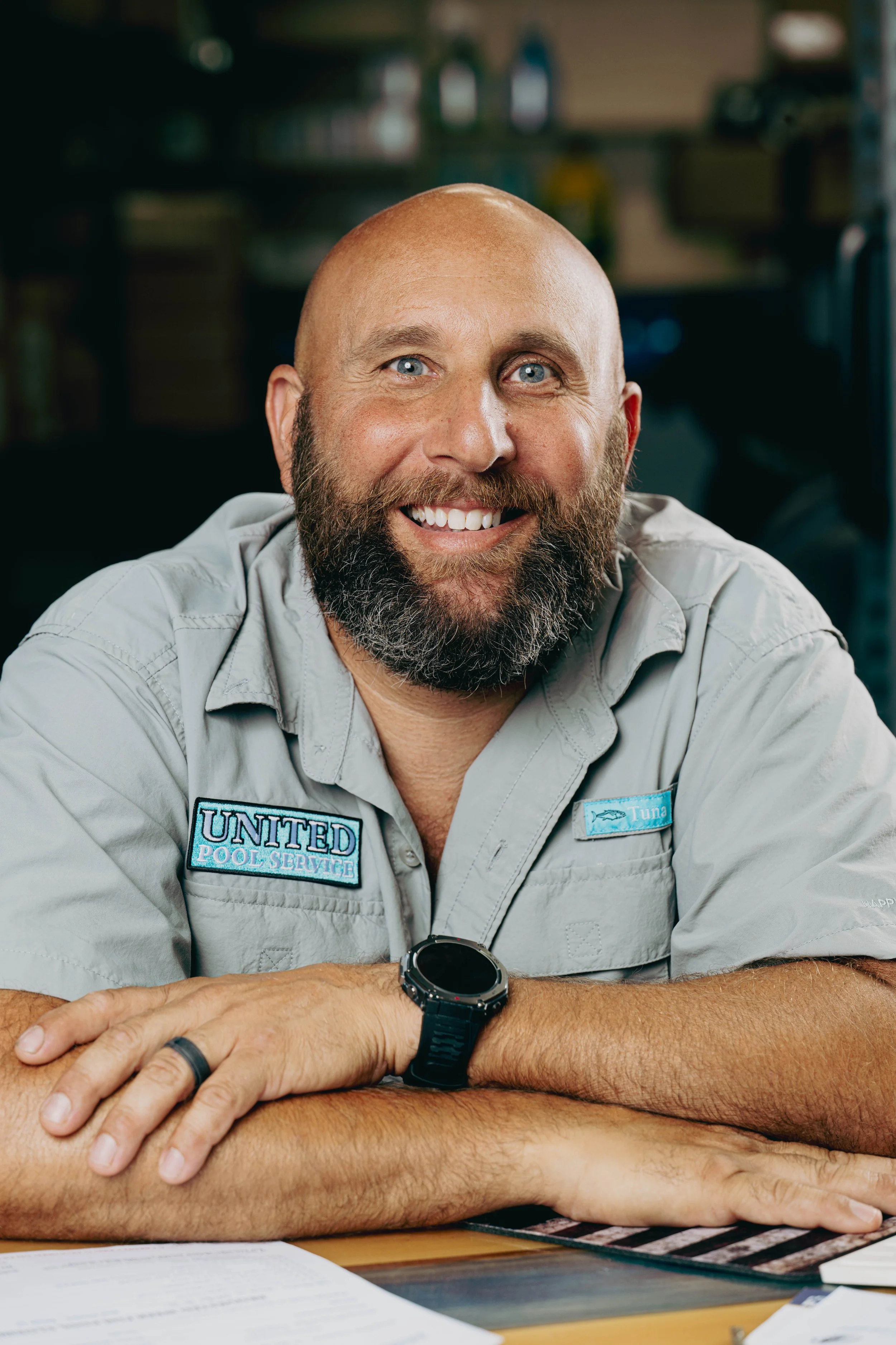 A smiling man with a beard, wearing a uniform with patches that read 'United Pool Service' and 'Tuna,' sitting at a table with papers in front of him.