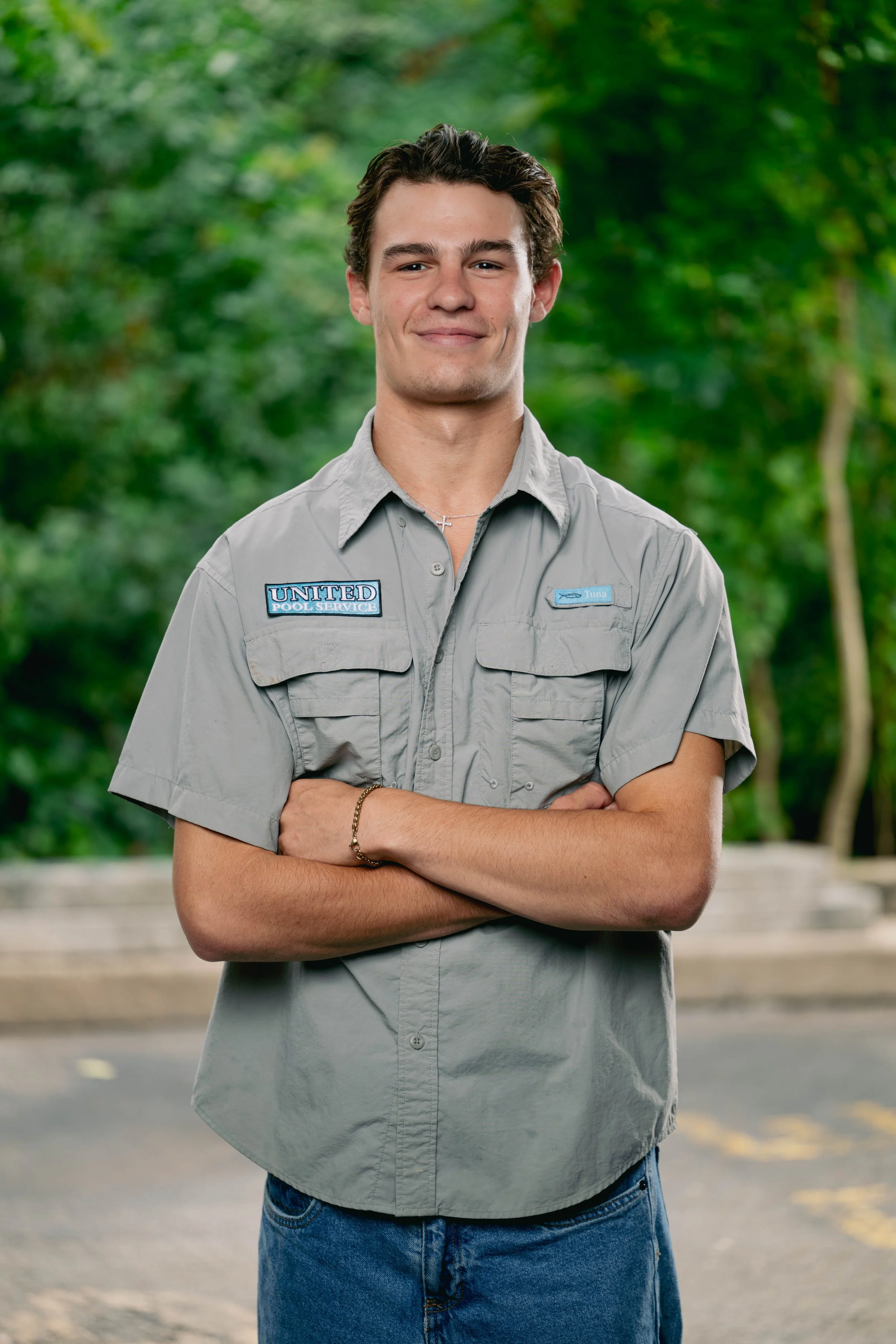 Young man with dark brown hair smiling, wearing a gray uniform with patches that say 'United Pool Service' and 'Luna,' standing outdoors with green trees in the background.
