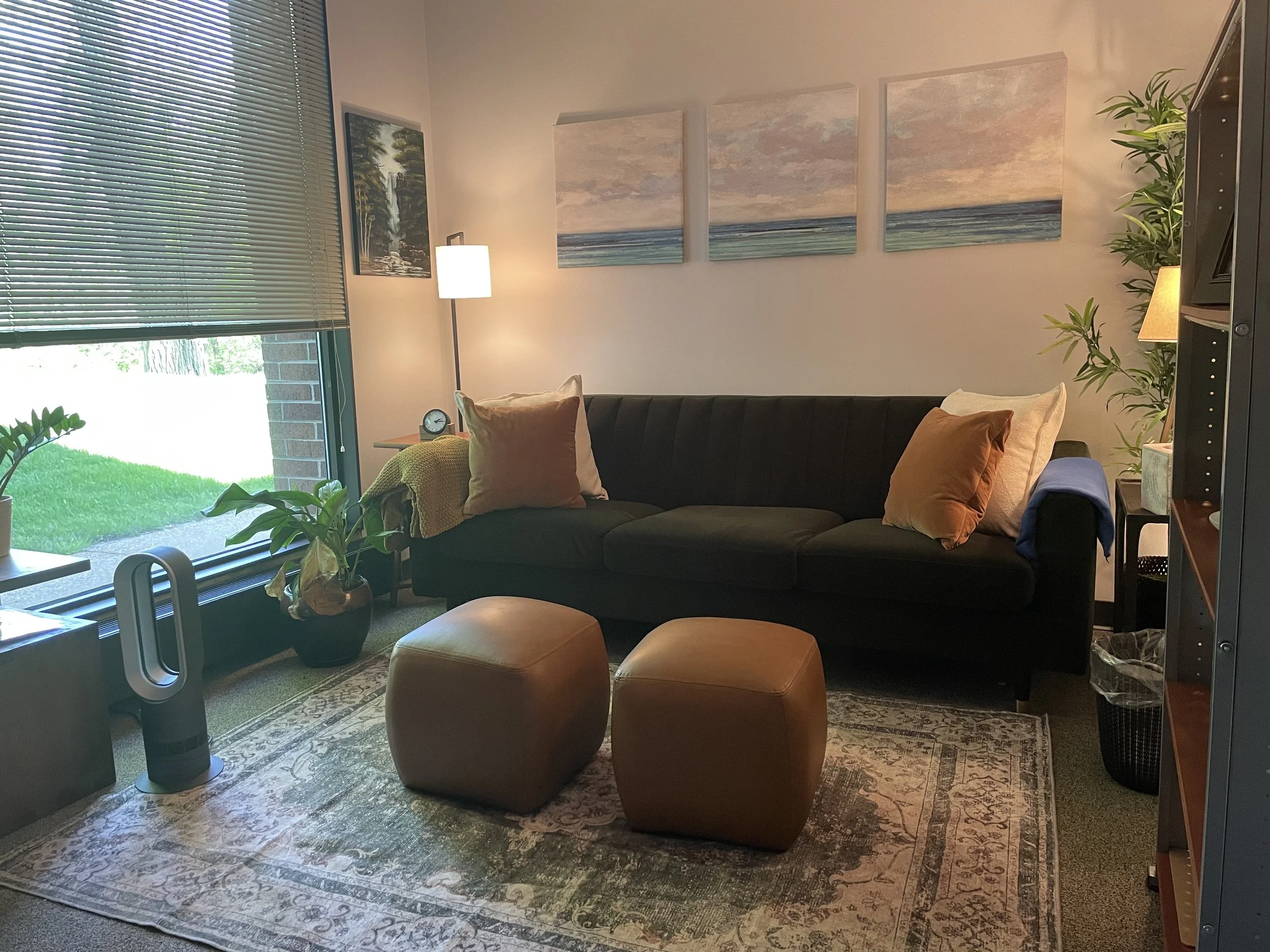 A cozy living room with a dark sofa, two caramel-colored ottomans, indoor plants, a lamp, and wall art of a beach scene. There is a large window with blinds, and outside green grass is visible.