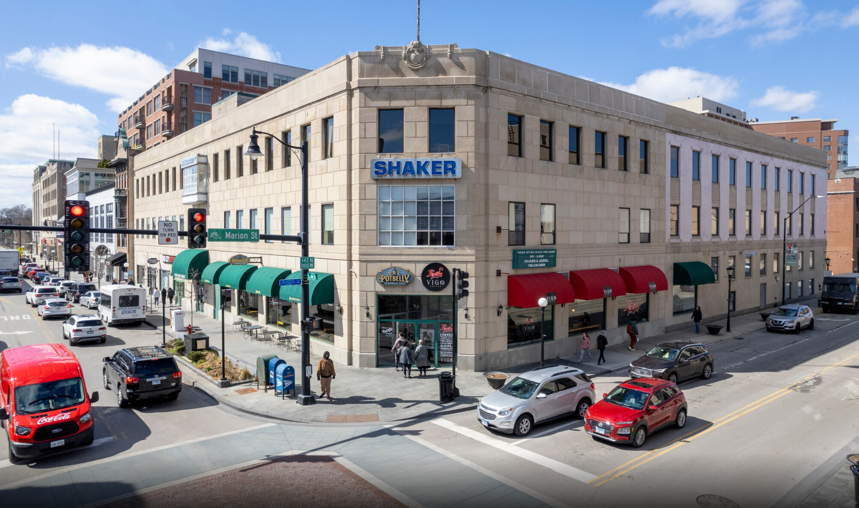 Street corner with a large beige building labeled 'SHAKER' and shops with green and red awnings, cars stopped at traffic lights, and pedestrians walking on the sidewalks.