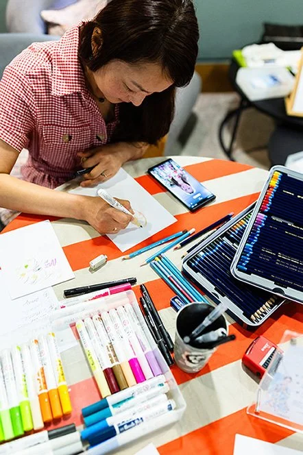 A photo of Live event illustrator Siya Liu drawing a lady on a table with maker pens and colour pencils.