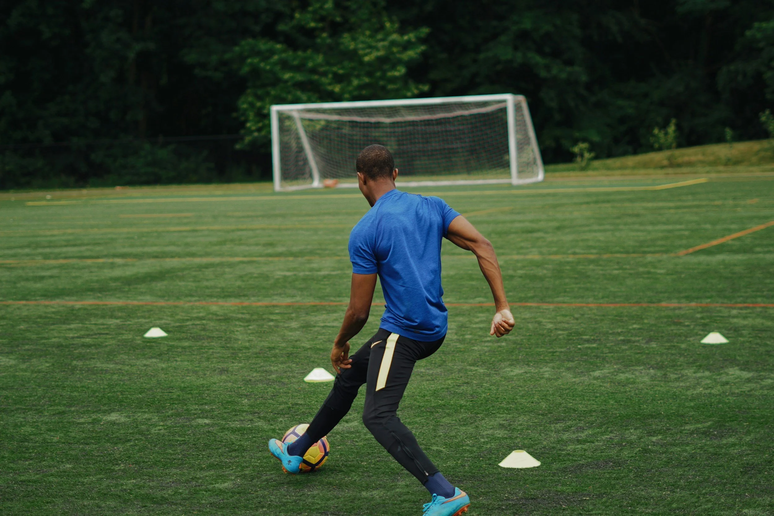 A man in a blue shirt and black pants practicing soccer on a perfectly cut grass field, surrounded by small white cones, with a soccer goal in the ready to be scored in.