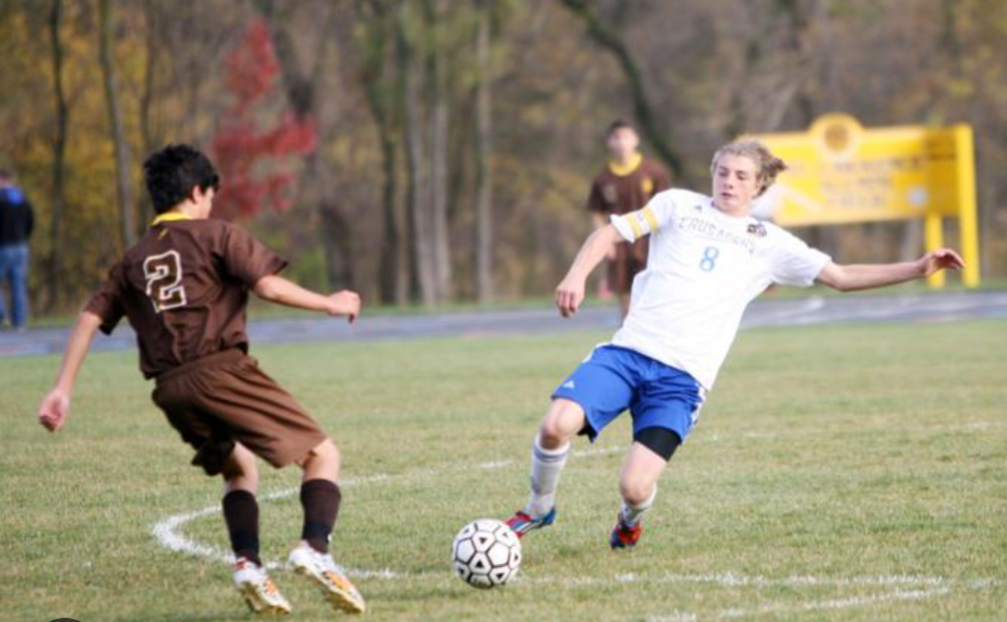 Two young soccer players are playing on a grassy field during the daytime. One player in a brown uniform is approaching the player in a white and blue uniform, who is kicking the soccer ball. Trees with autumn foliage are in the background.
