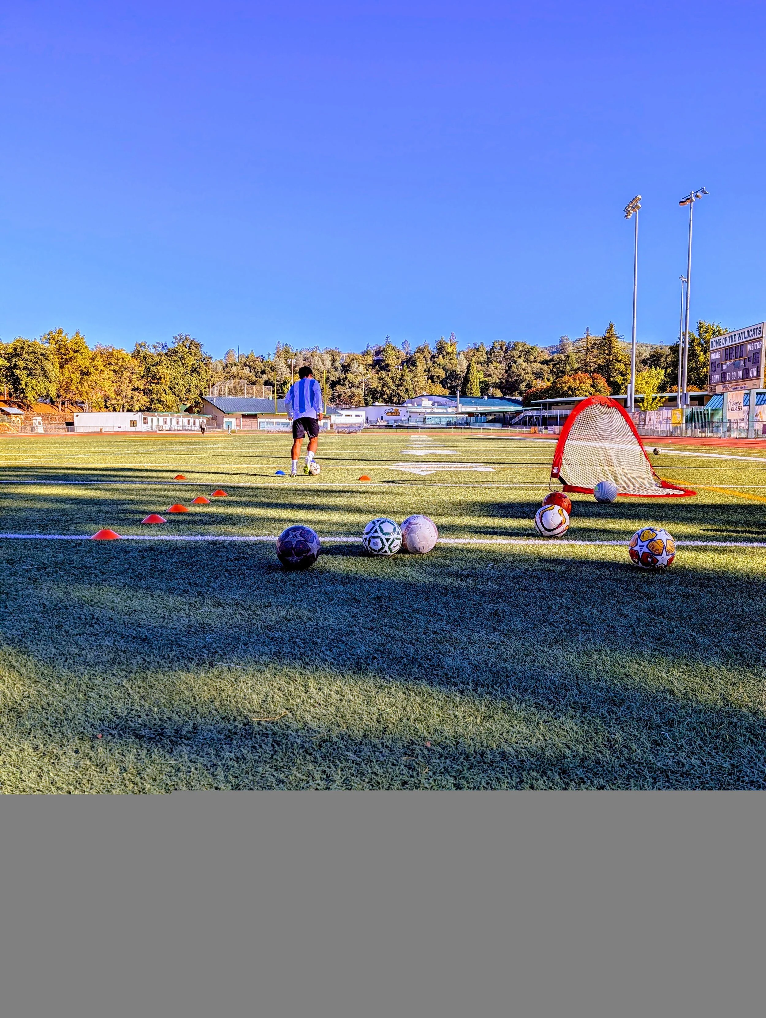 A person practicing soccer on a field with multiple soccer balls, a small goal, orange cones, and a track, under a clear blue sky.