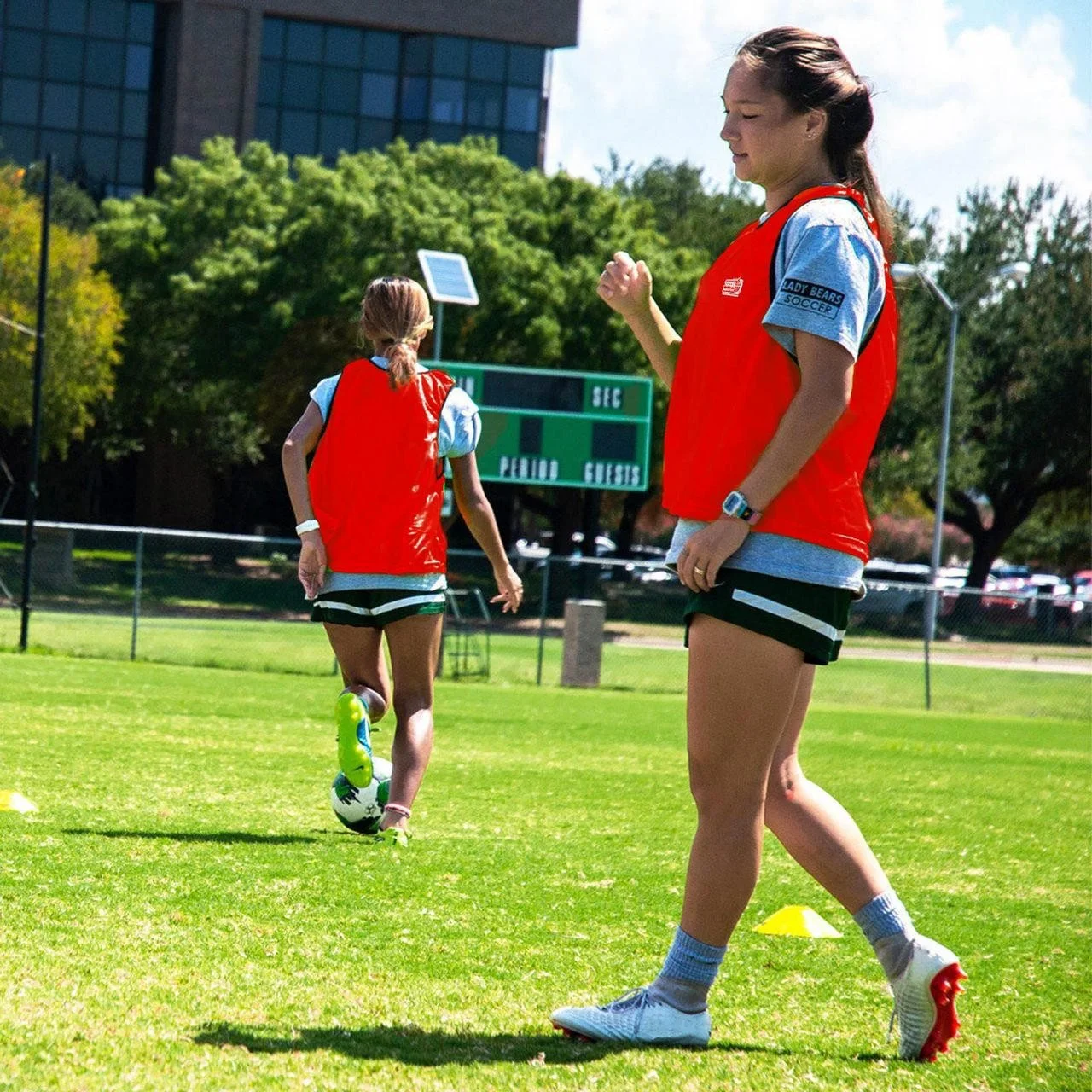 Soccer players competing on a green field with spectators and parked cars in the background during daytime.