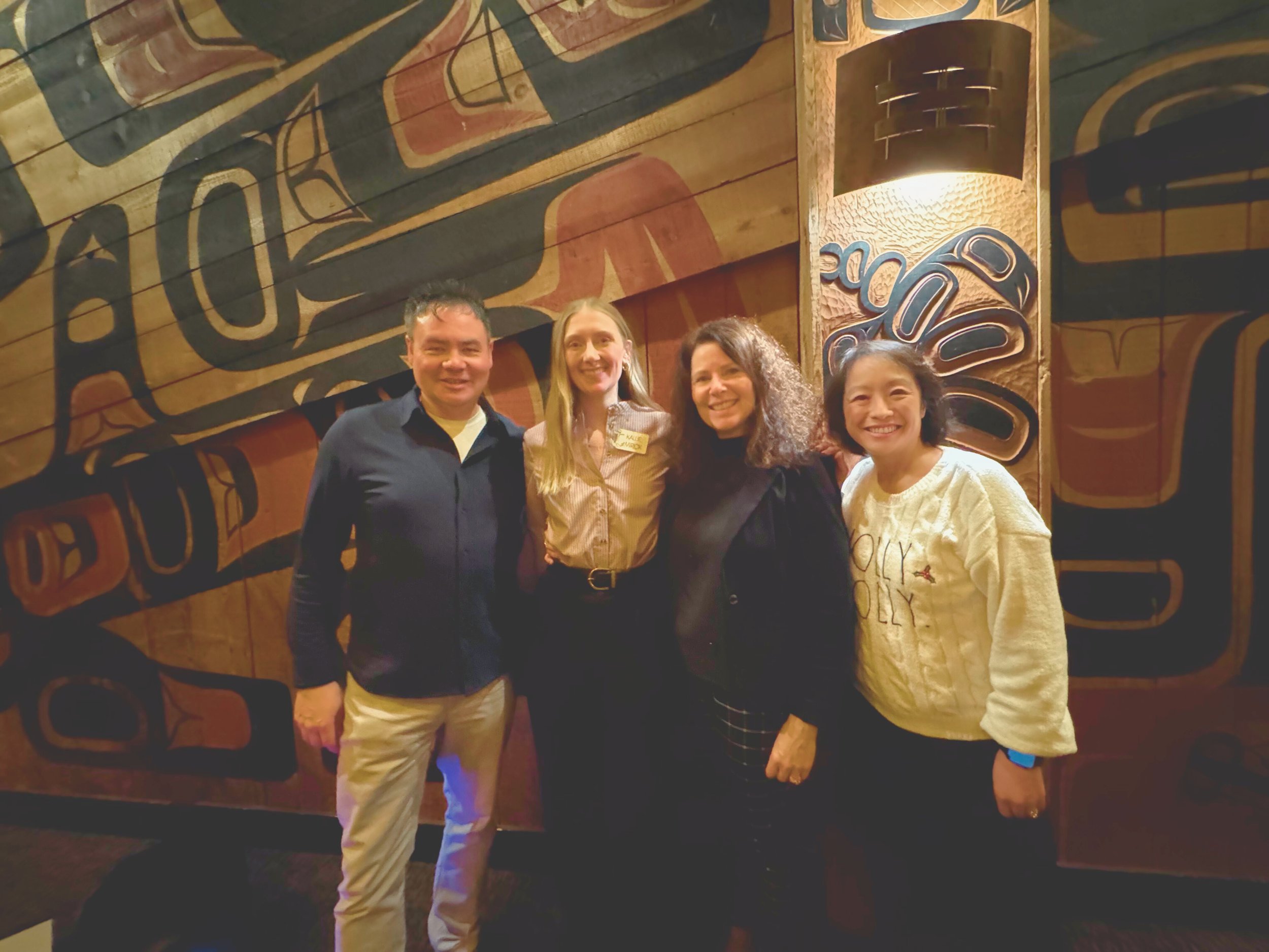 Four smiling people standing together in front of a colorful wooden wall with tribal-style artwork.