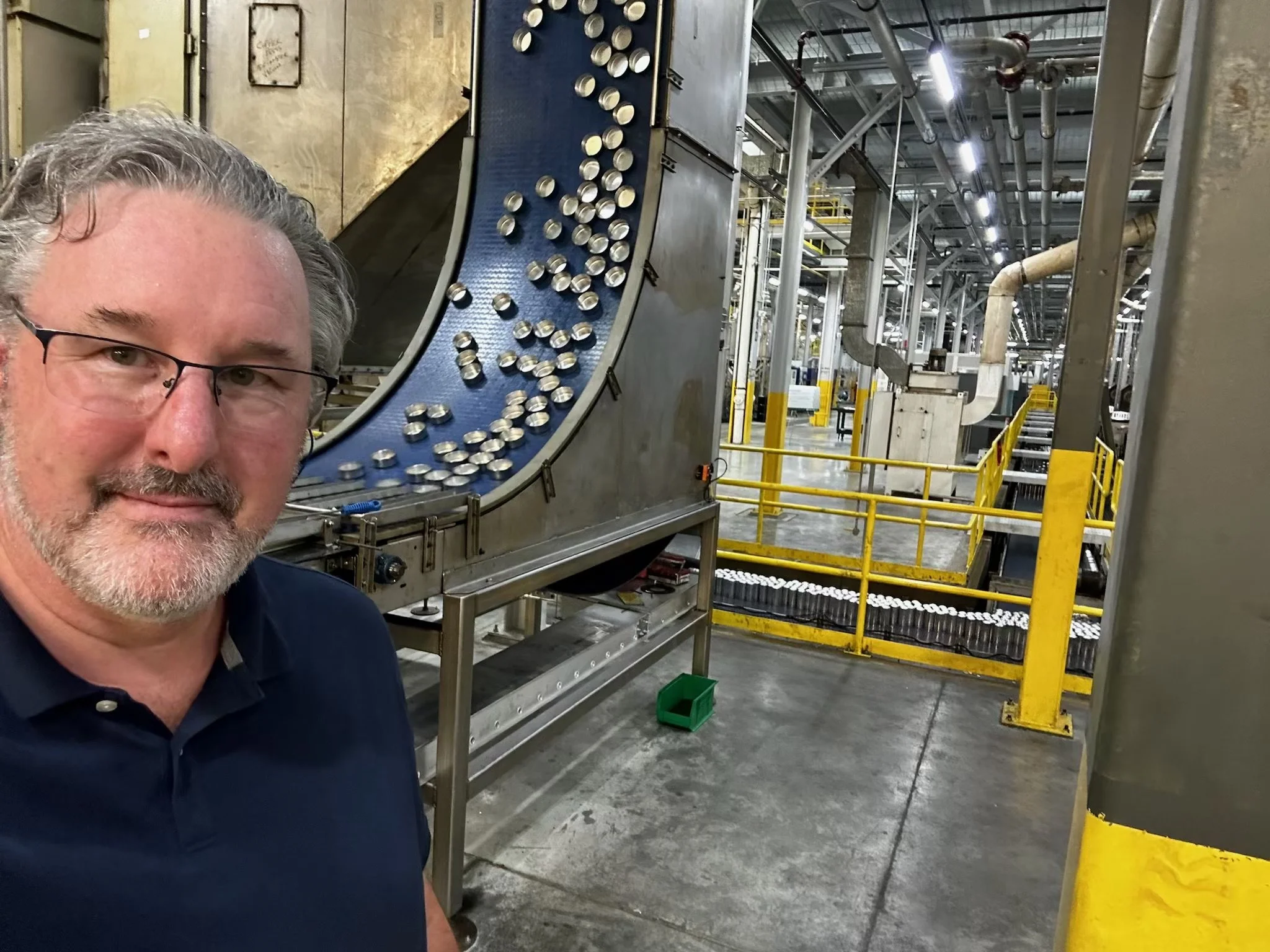 A man with glasses and gray hair taking a selfie inside an industrial facility with machinery and conveyors in the background.