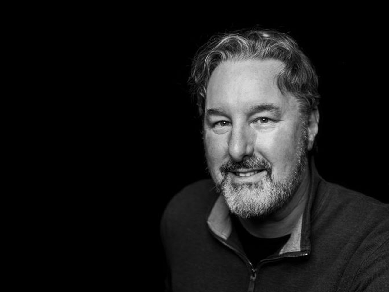 Black and white portrait of a smiling man with wavy hair and a beard, wearing a zip-up sweater, against a dark background.