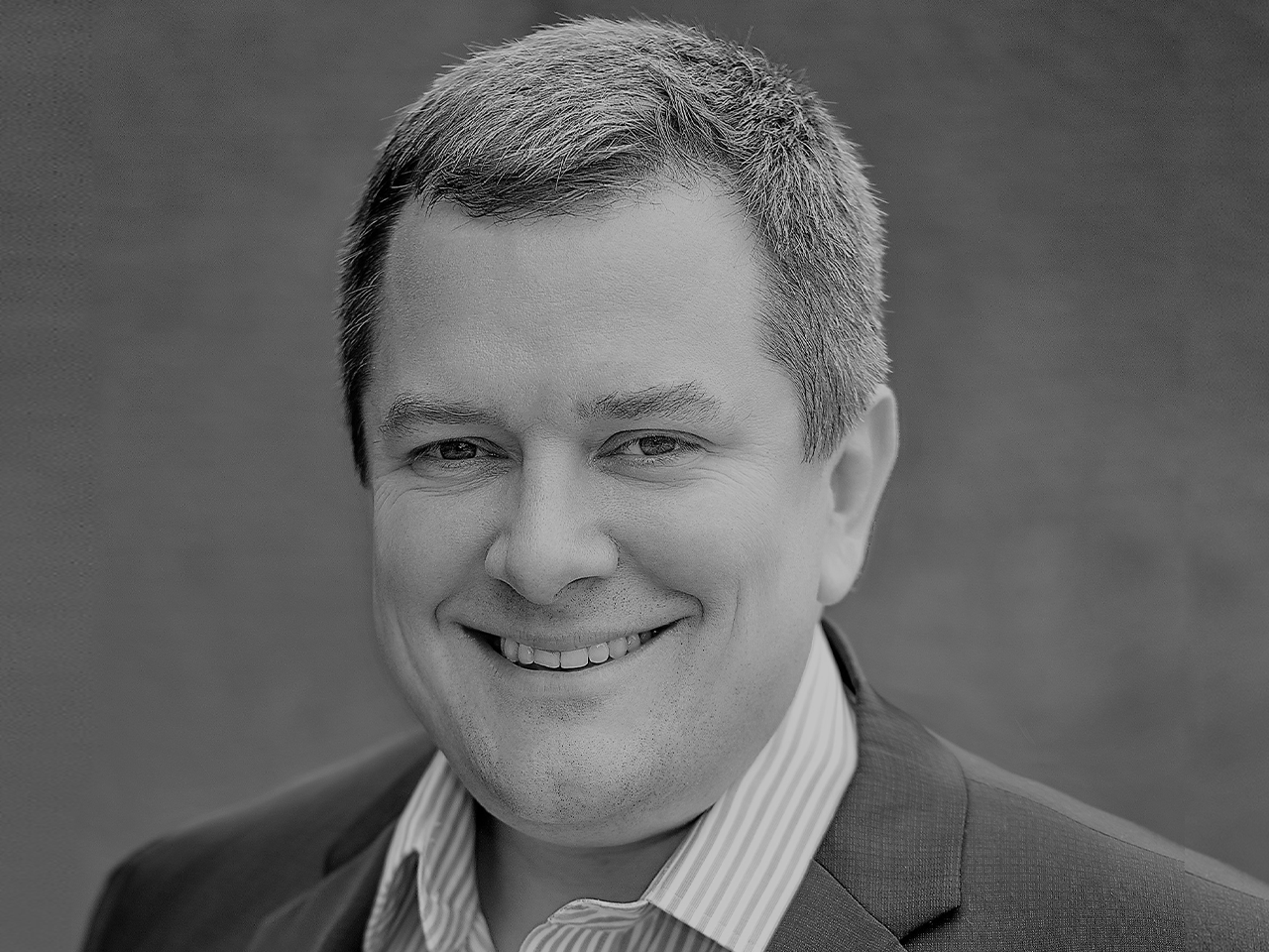 Black and white headshot of a smiling man in a suit and striped shirt.