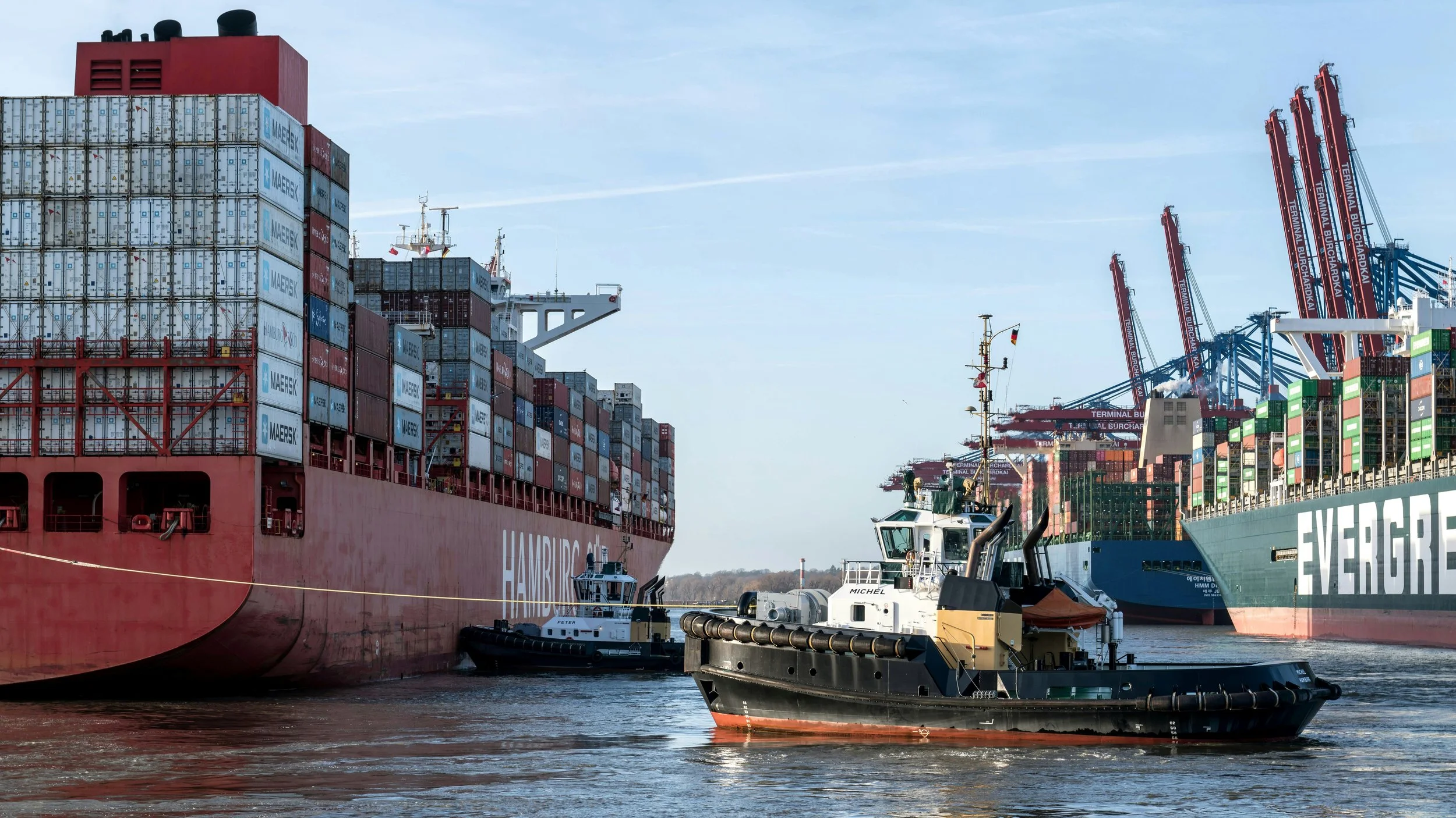 Cargo ships with containers docked at a port, with a tugboat in the foreground, cranes in the background, and a clear sky.
