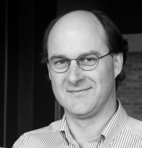 Black and white portrait of a man with glasses, receding hairline, wearing a striped shirt, smiling at the camera.
