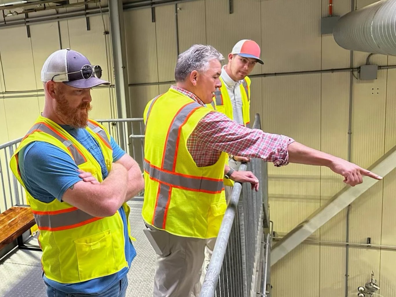 Three men in yellow safety vests standing on a metal platform, looking and pointing towards an area below inside an industrial or warehouse setting.