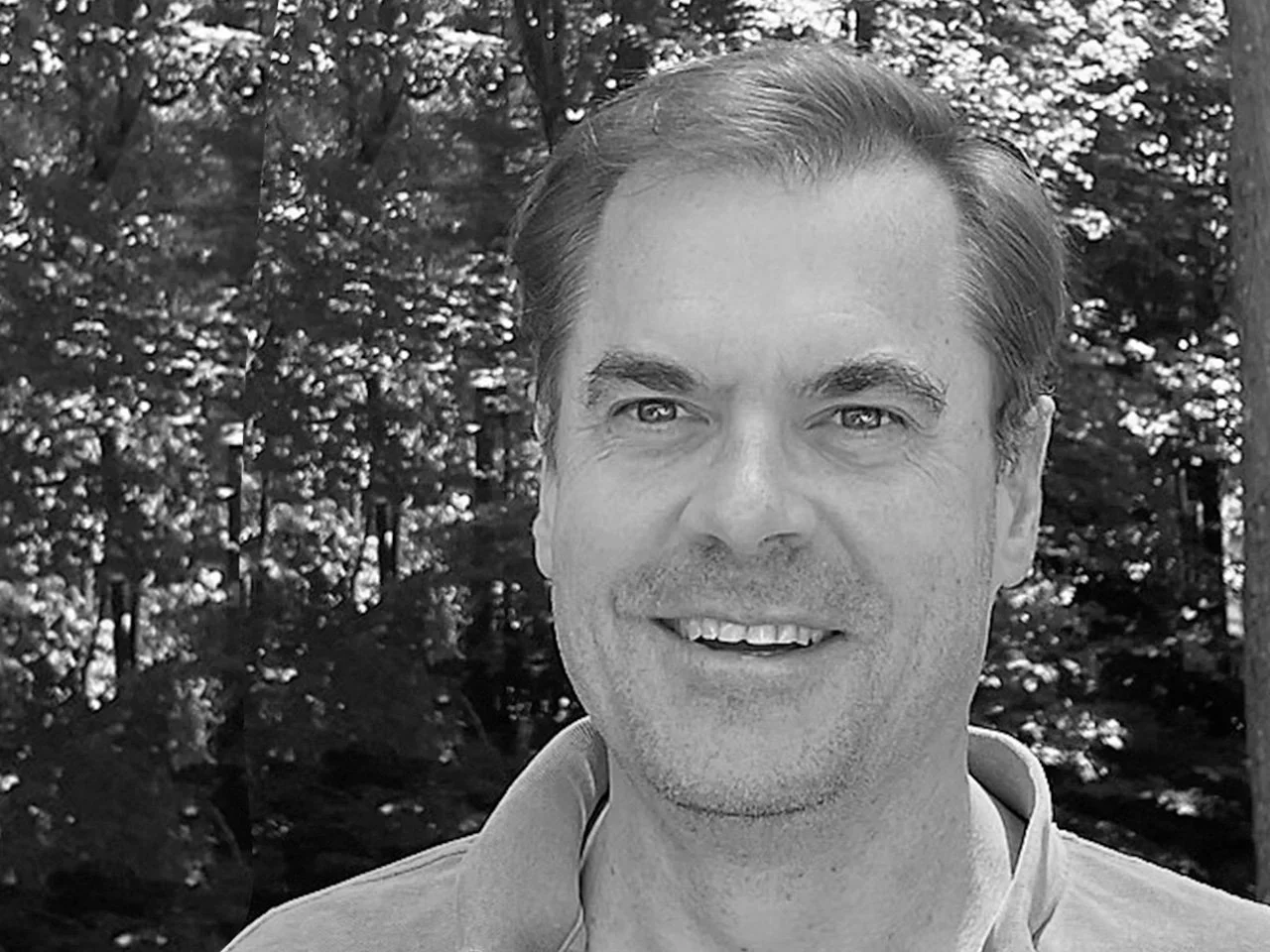 Black and white photo of a man with short hair and a beard smiling outdoors, with trees in the background.