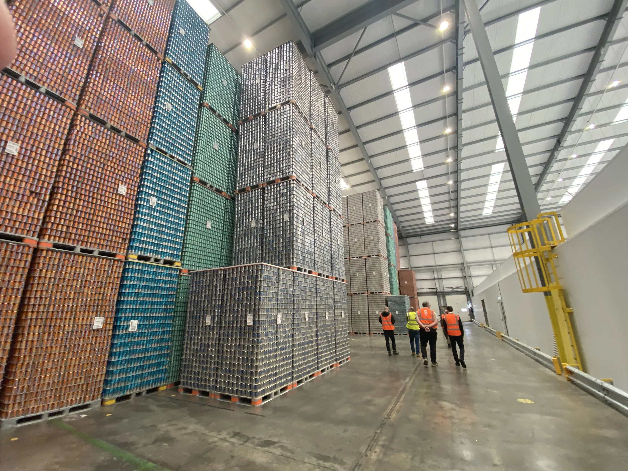 Warehouse with stacks of soda cans organized by color, four workers in safety vests walking through the aisle.