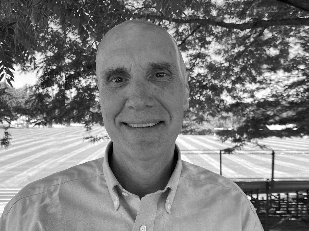 Black and white photo of a smiling man wearing a collared shirt, standing outdoors with trees and a structure in the background.