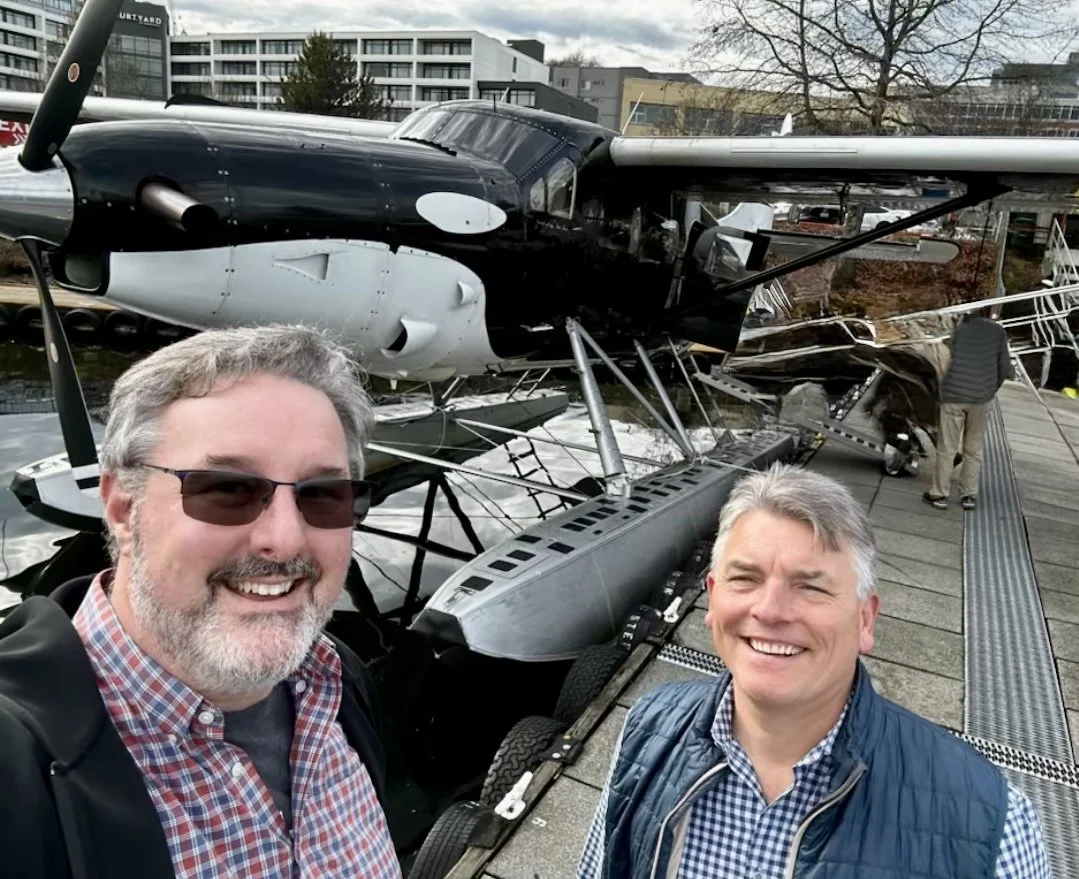 Two men smiling in front of a small aircraft at an outdoor location.