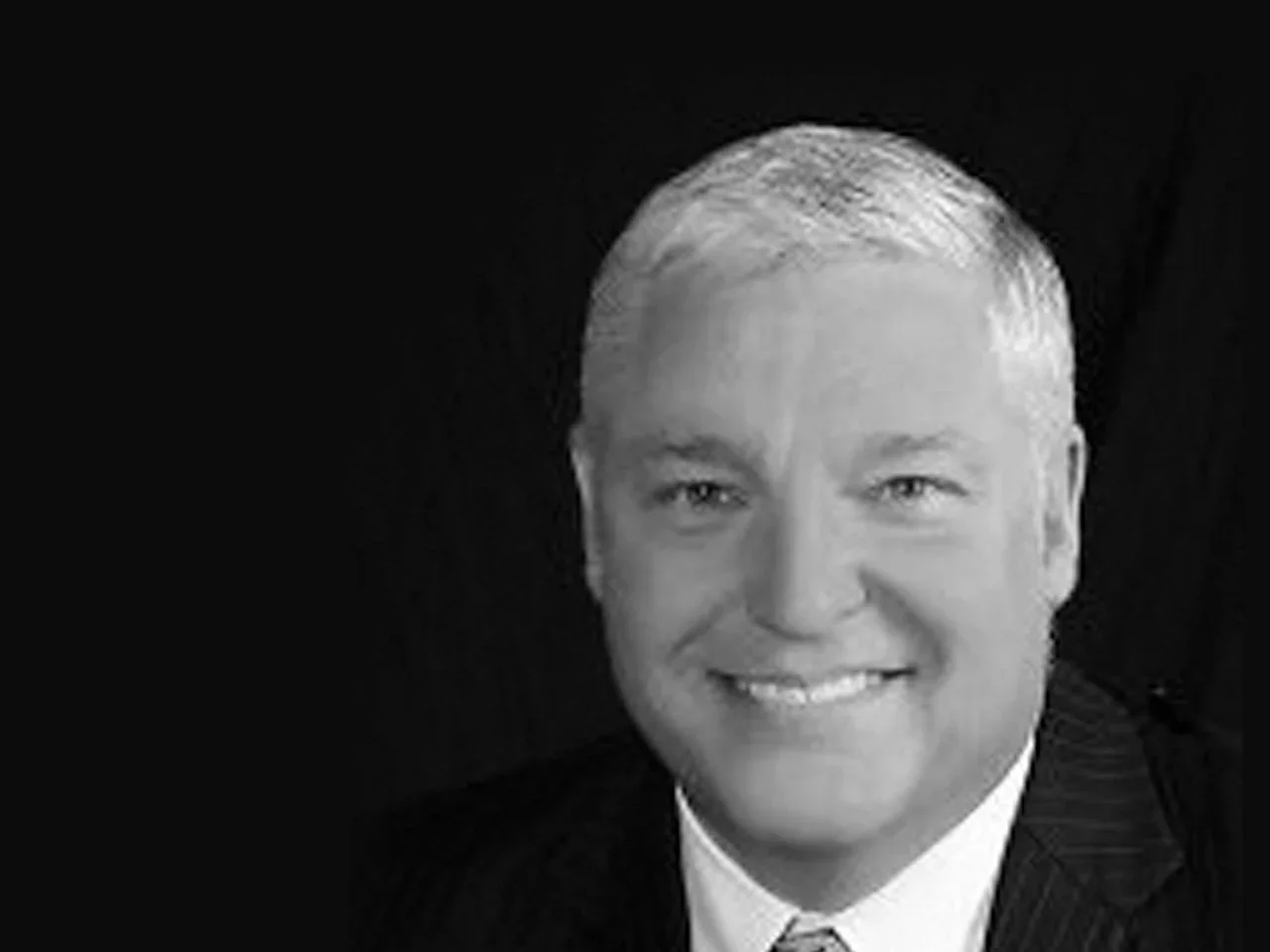 Black and white photo of a smiling man with short, neatly combed hair, wearing a suit and tie.