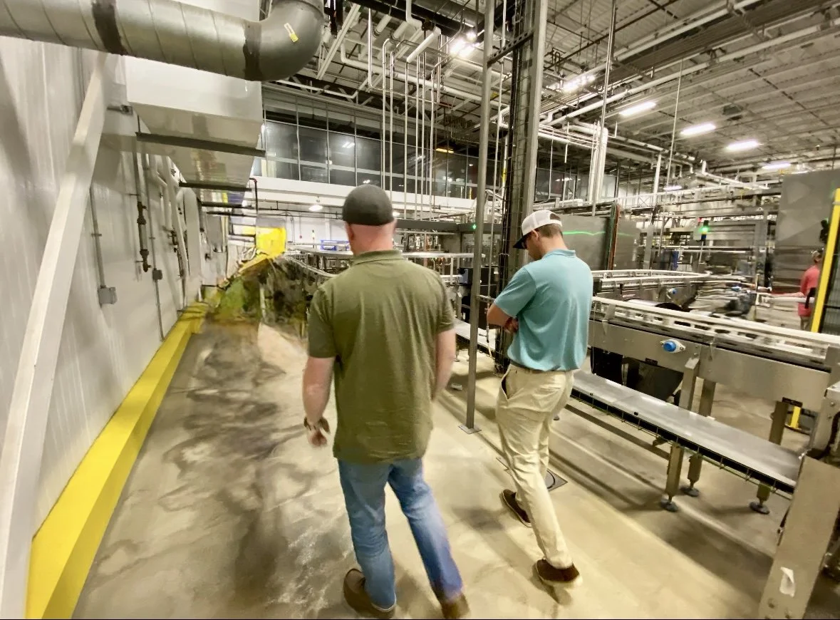 Two people walking through an industrial facility or factory, surrounded by machinery, piping, and conveyor belts.