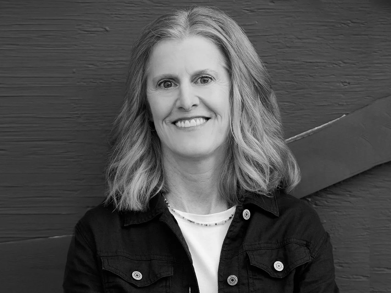 Black and white portrait of a smiling woman with shoulder-length wavy hair, wearing a dark jacket over a light shirt, standing in front of a wooden background.