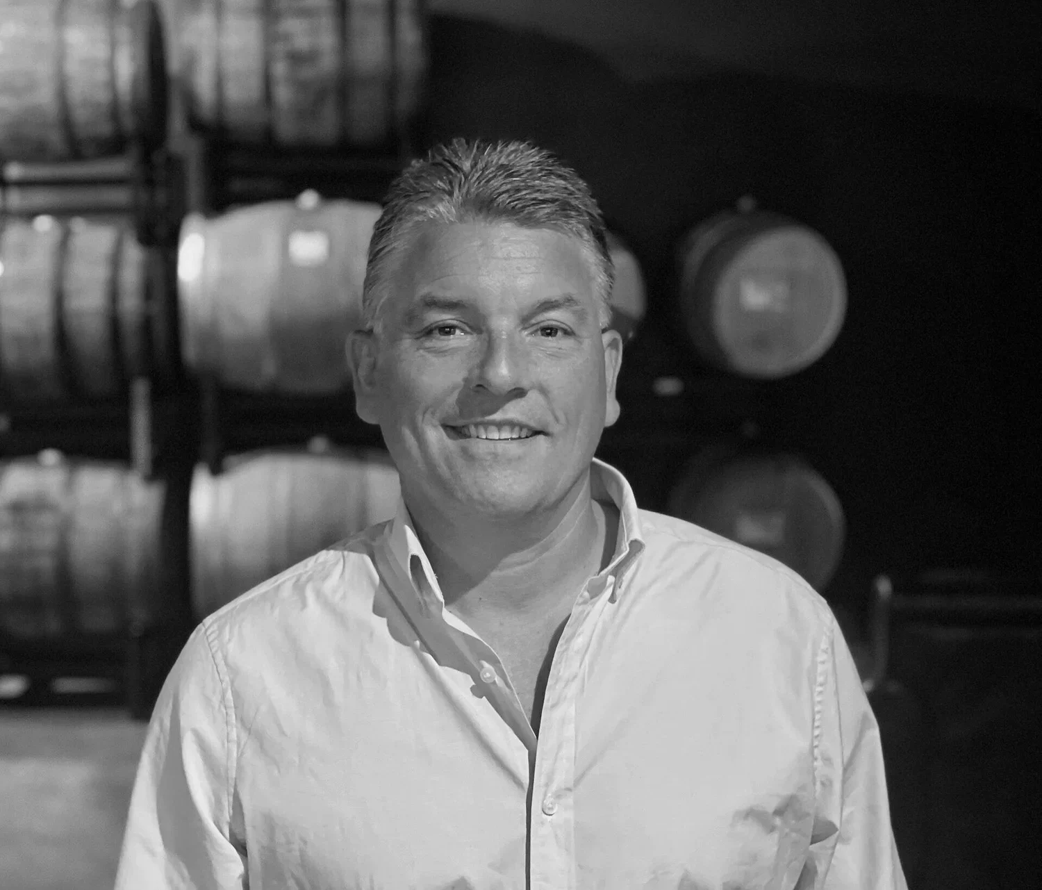 A smiling man in a white shirt standing in front of stacked wine barrels in a cellar.