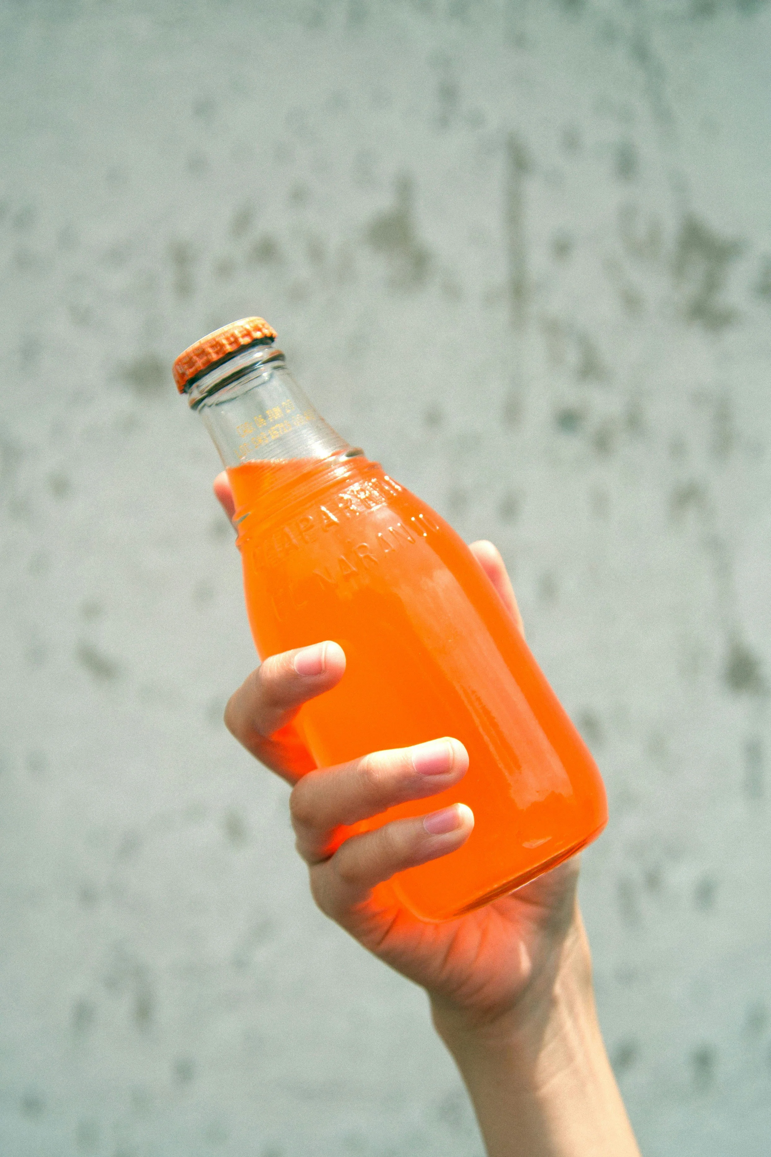 A hand holding a glass bottle with bright orange liquid against a blurred light gray background.