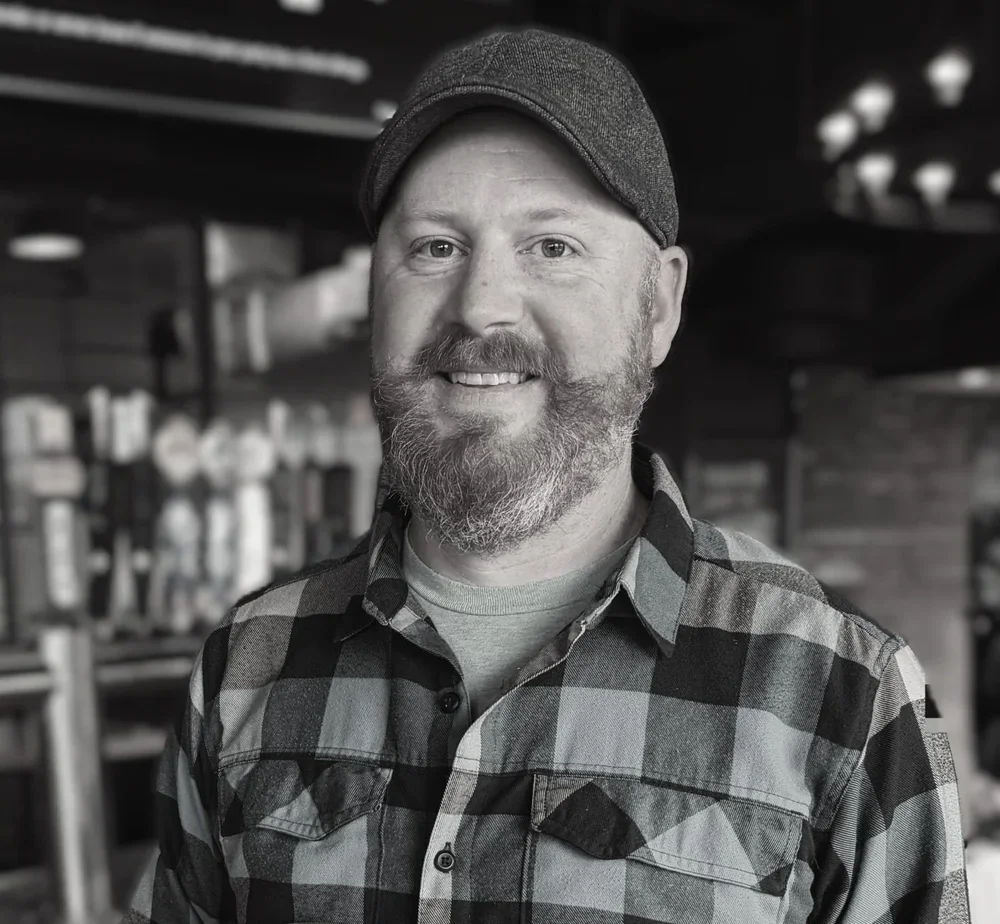 A man with a beard and mustache wearing a checkered shirt and a baseball cap, smiling in an indoor setting.