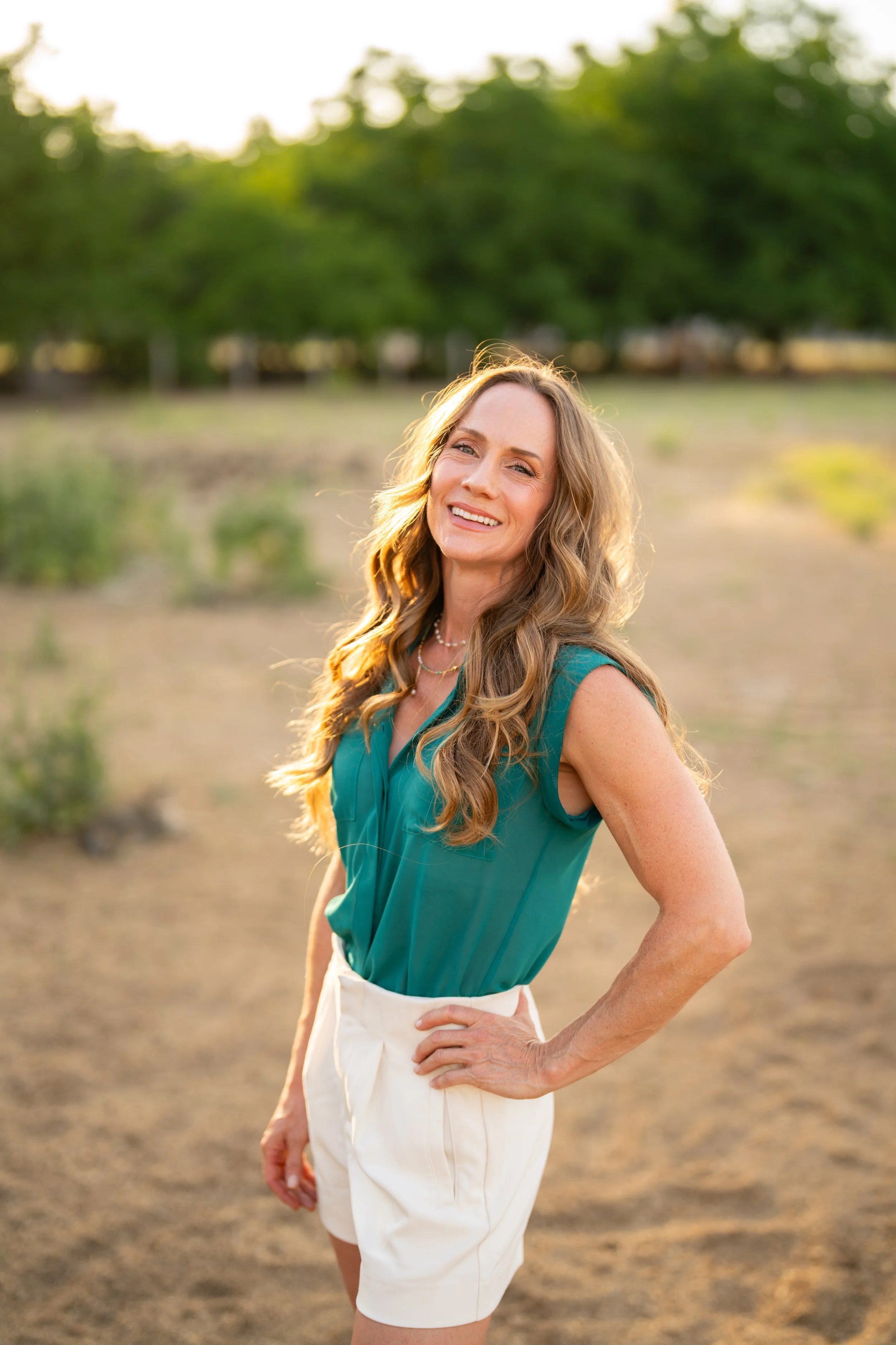 A smiling woman with long wavy hair, wearing a teal sleeveless blouse and white high-waisted pants, standing outdoors in a sunny field with green trees in the background.