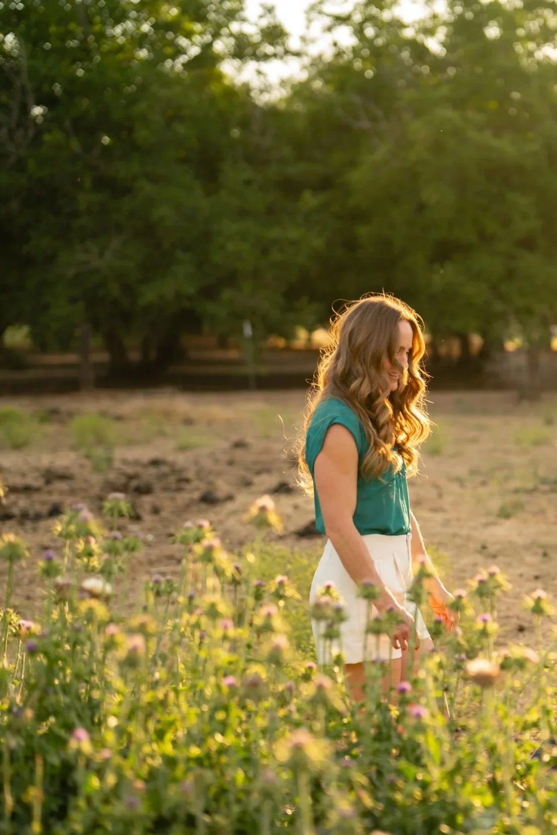 A woman with long wavy hair wearing a teal top and white shorts standing in a field of pink and yellow flowers, with trees and sunlight in the background.