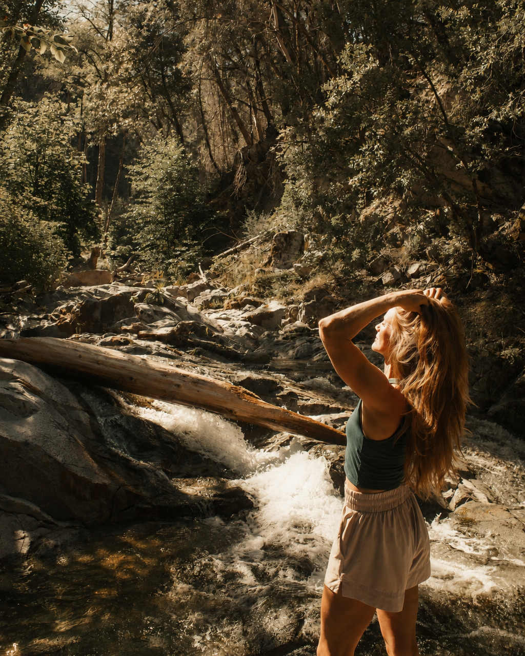 A woman with long, wavy hair standing in a river in a forest, looking up while holding her head with one hand, surrounded by trees and rocks, during sunlight.