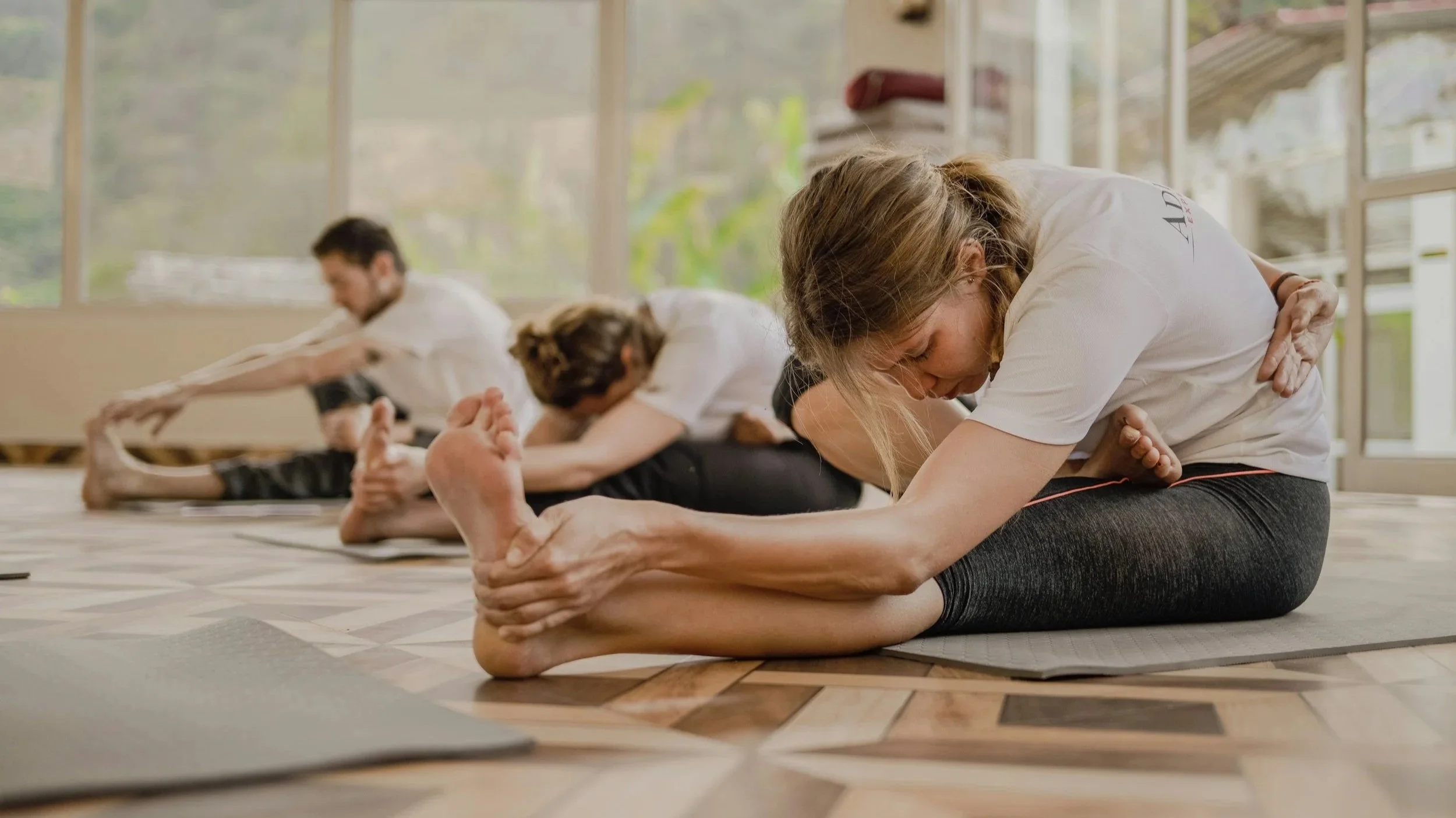 Group of people practicing yoga in a brightly lit indoor studio, performing seated forward bend stretches on yoga mats.