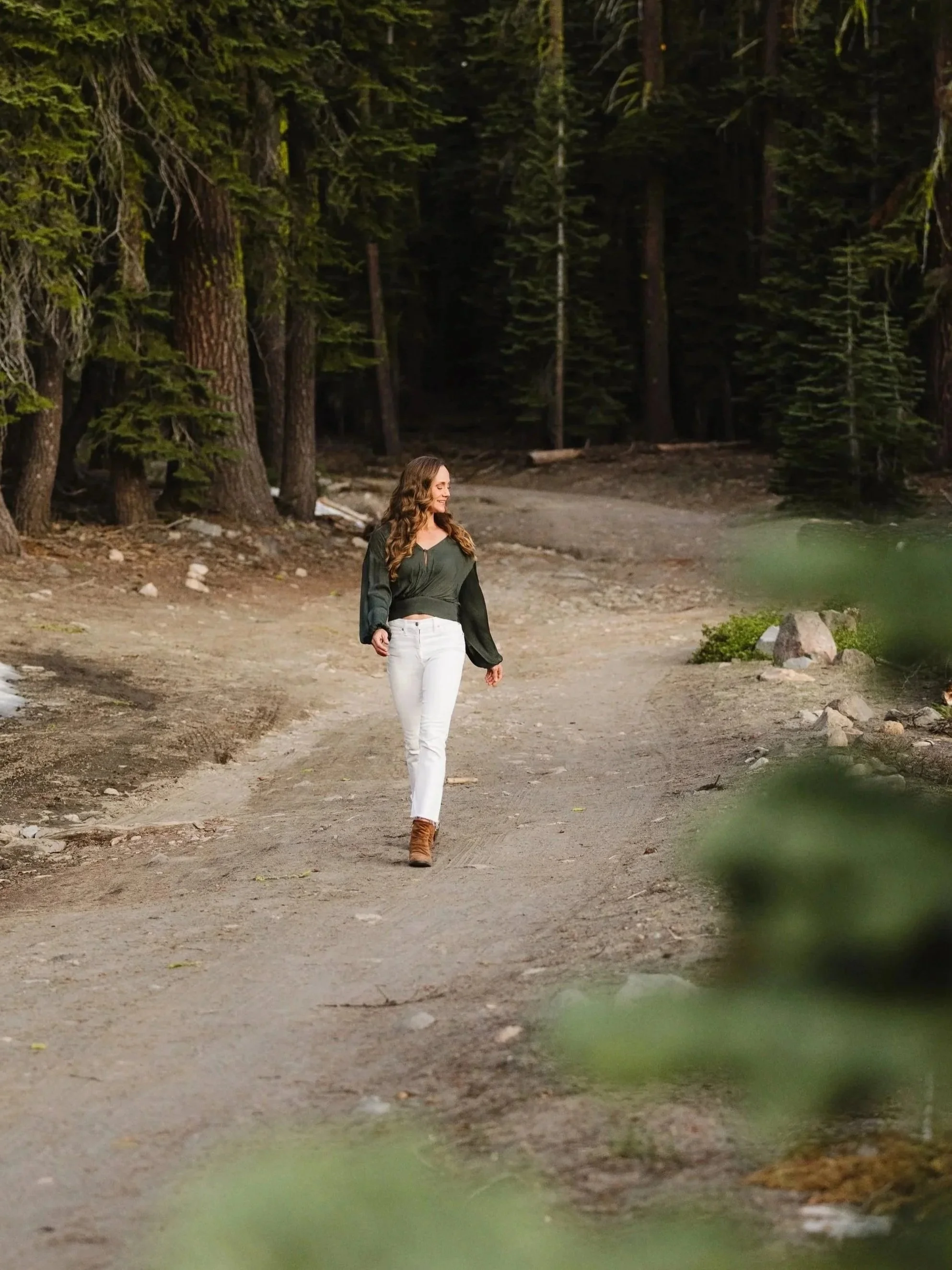 A woman with long, curly hair wearing a dark green blouse, white pants, and brown boots walking on a dirt path in a forest with tall trees and dense foliage.