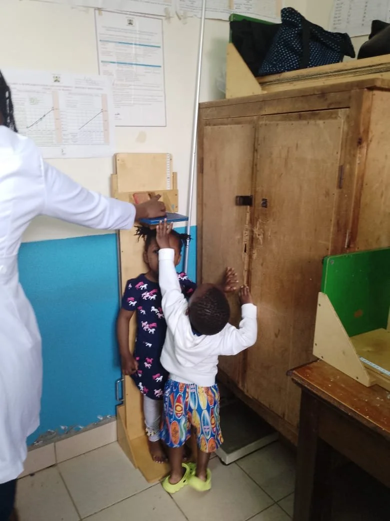 Two children are playing with a wooden cabinet in a room, while an adult observes. The girl is standing on a platform and holding the top of the cabinet, and the boy is reaching up towards the same cabinet. The room has posters on the wall and variou