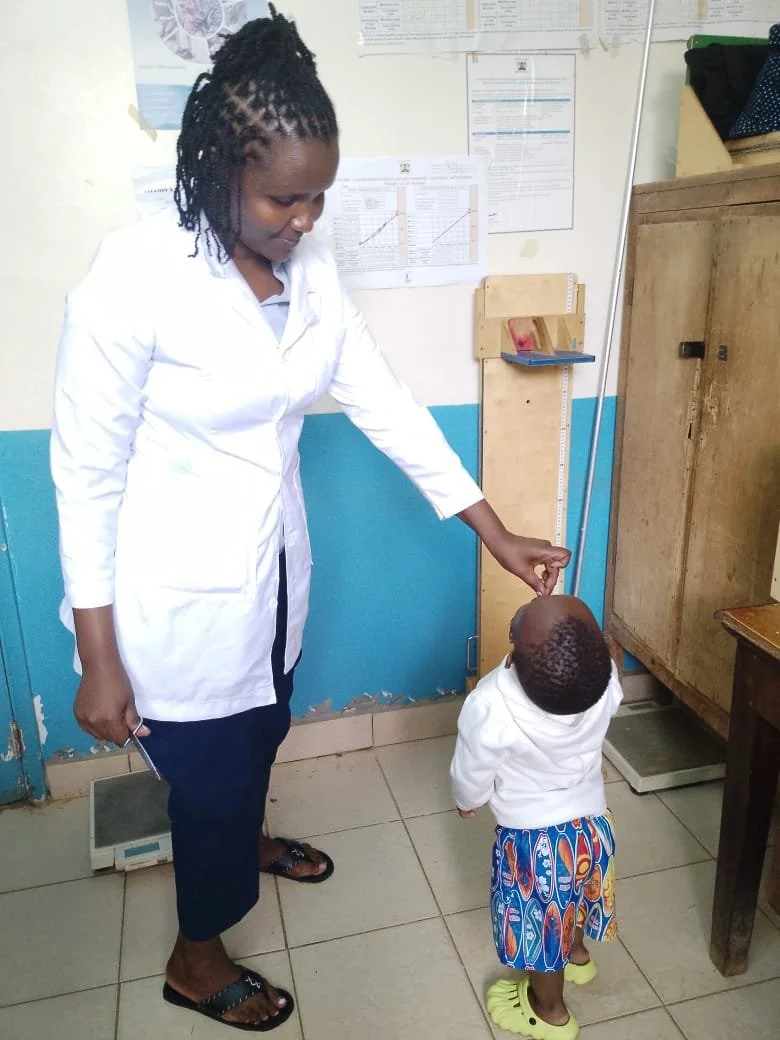 A woman in a white lab coat and dark pants is gently lifting the chin of a young child in a white hoodie and colorful shorts, inside a room with blue and white painted walls, posters, and a wooden cabinet.